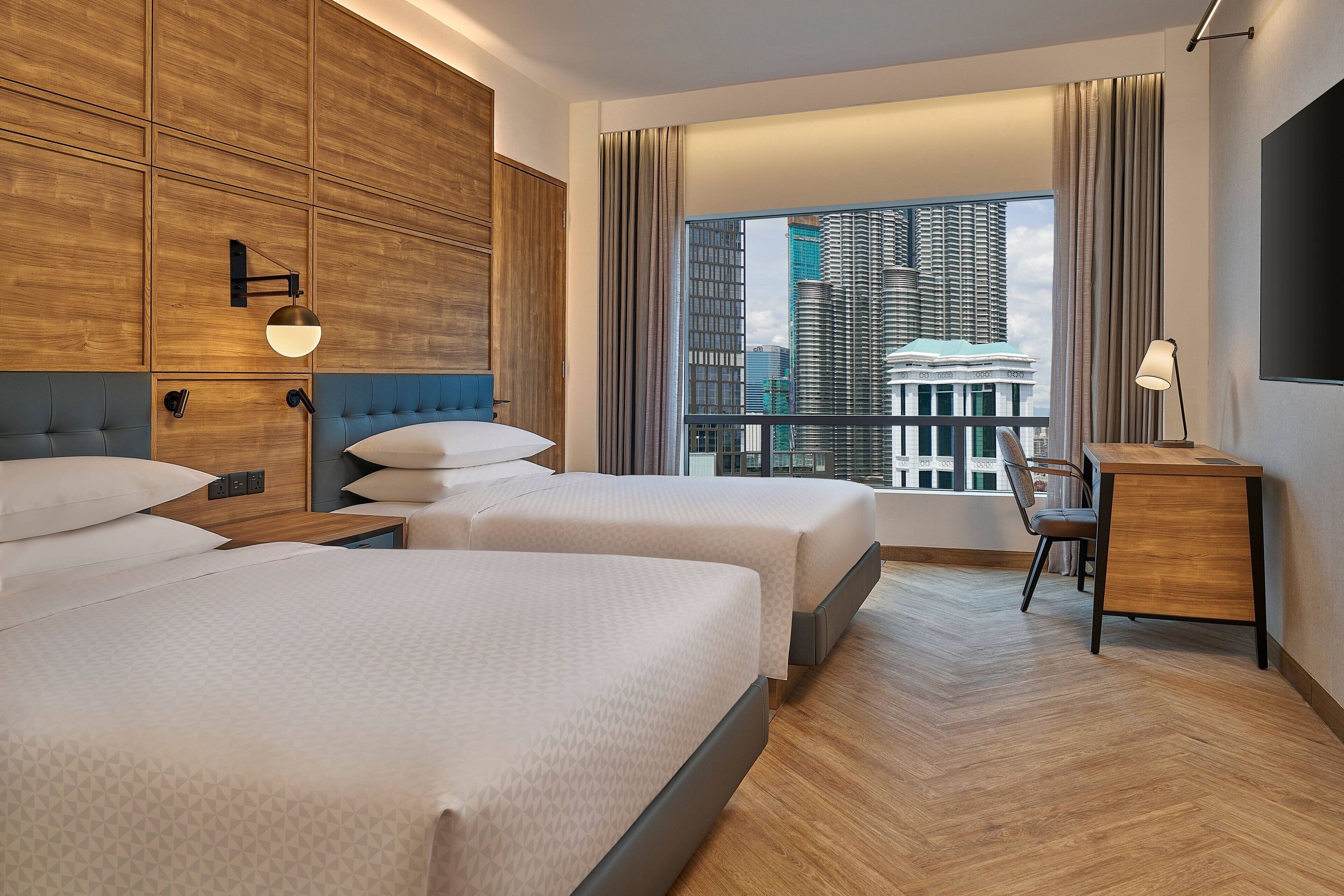 A hotel room with two beds, a desk, a chair, a large window with a city view, and a TV on the wall.