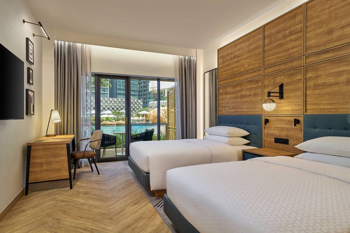Een hotelkamer met twee bedden, een bureau, een stoel en een groot raam met uitzicht op een zwembad en terras.