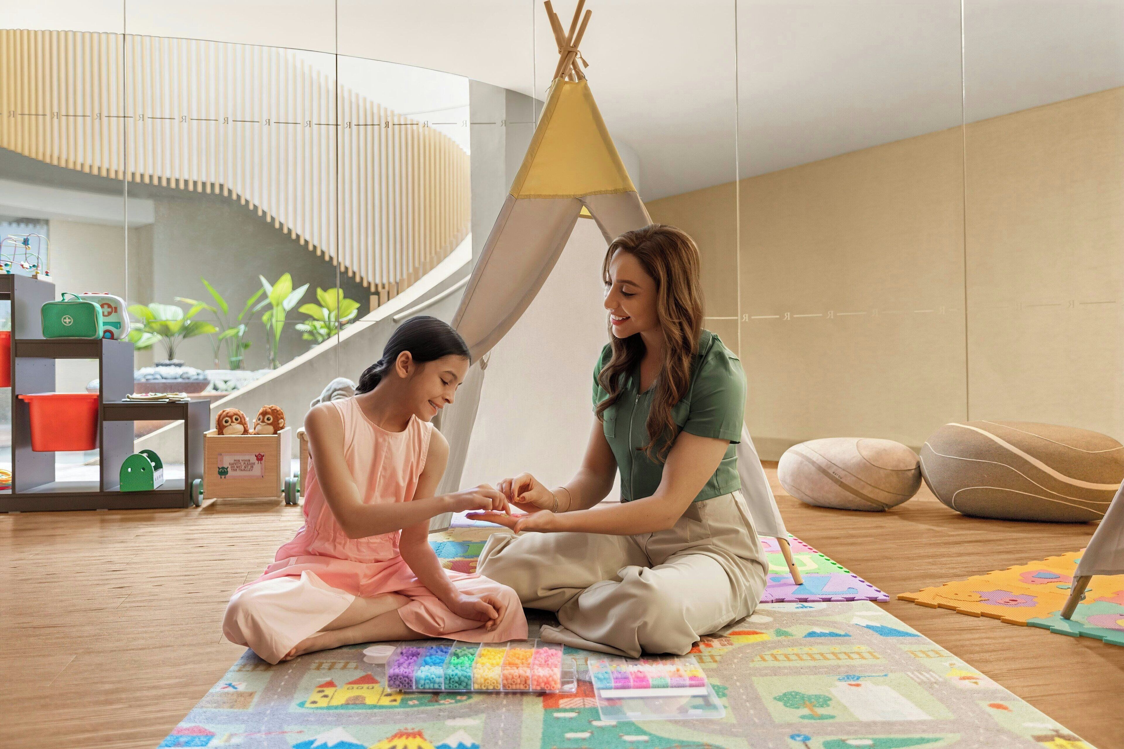 A room with a modern interior, a child and an adult sitting on a colorful play mat, a small tent-like structure, and a staircase in the background.