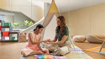 A room with a modern interior, a child and an adult sitting on a colorful play mat, a small tent-like structure, and a staircase in the background.