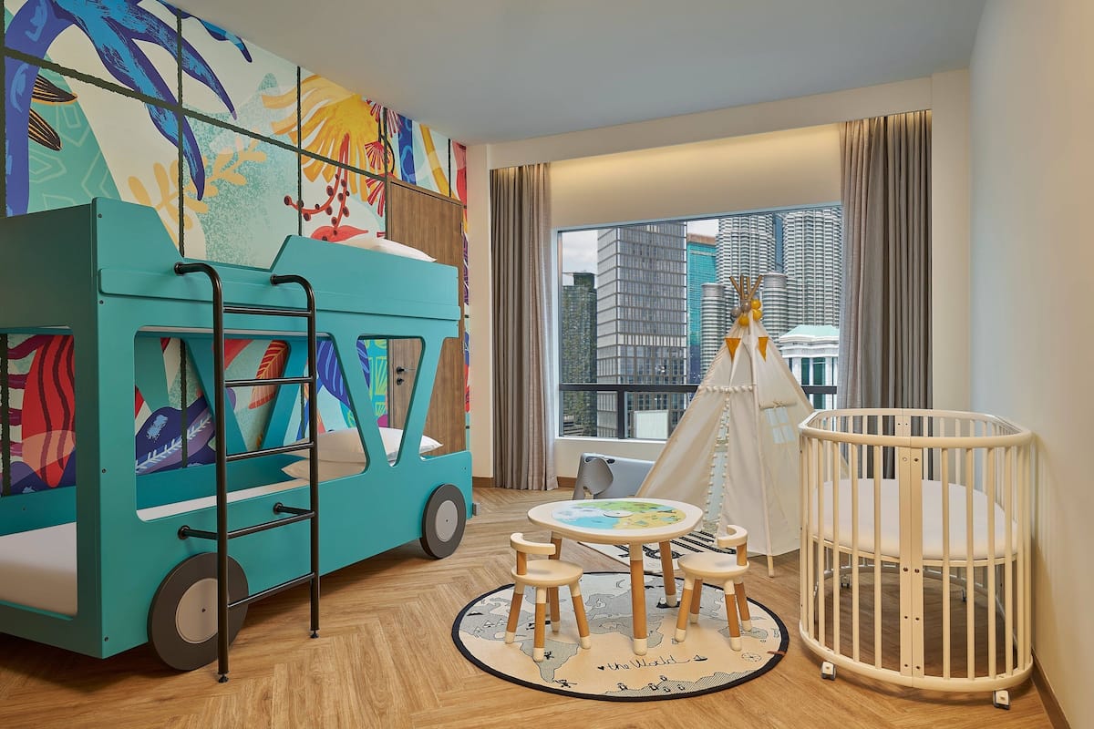 Een kinderspeelkamer met een kleurrijke speelauto, een klein rond tafeltje met krukjes, een wit ledikant en een tent.