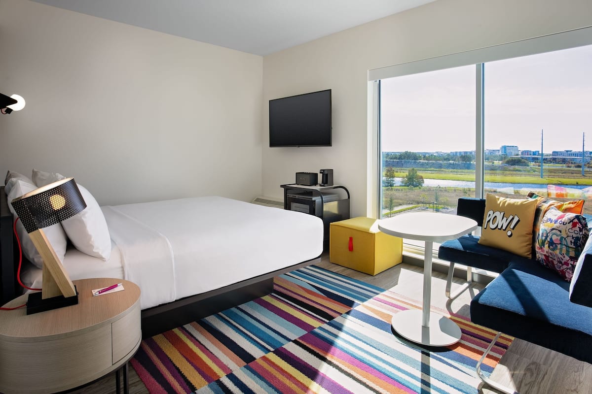 A hotel room with a bed, a TV, a desk with a lamp, a sofa, a small table, and a colorful striped rug.