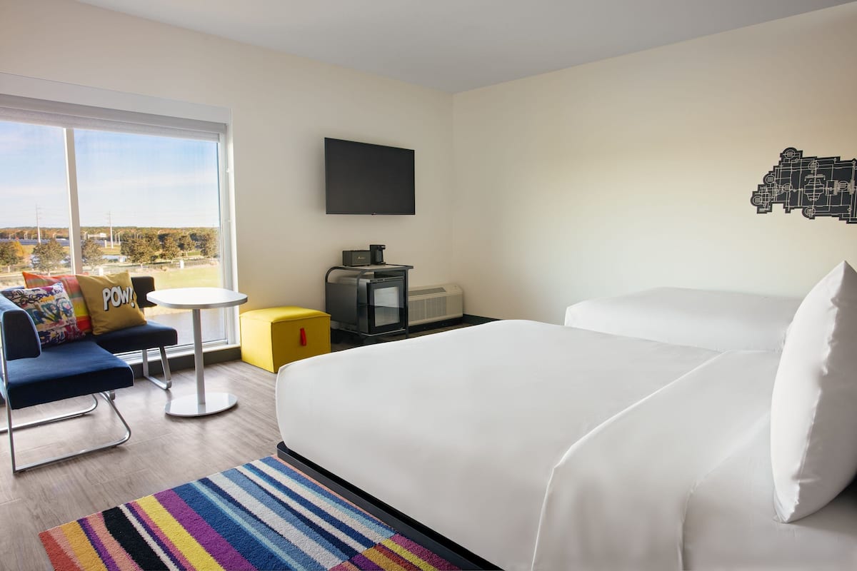 A hotel room with a large bed, a TV mounted on the wall, a small table, and a sofa with a colorful rug.