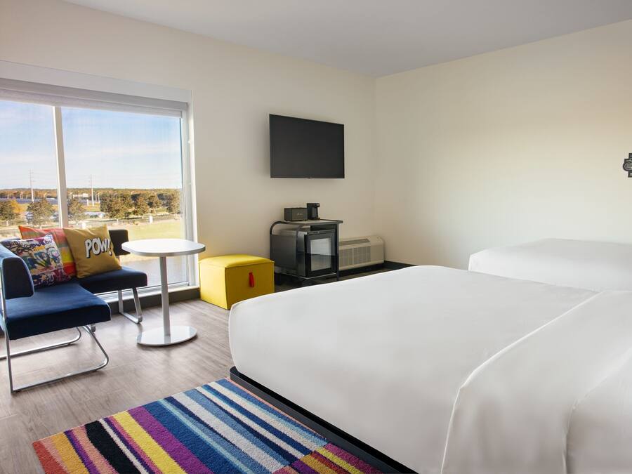 A hotel room with a large bed, a TV mounted on the wall, a small table, and a sofa with a colorful rug.