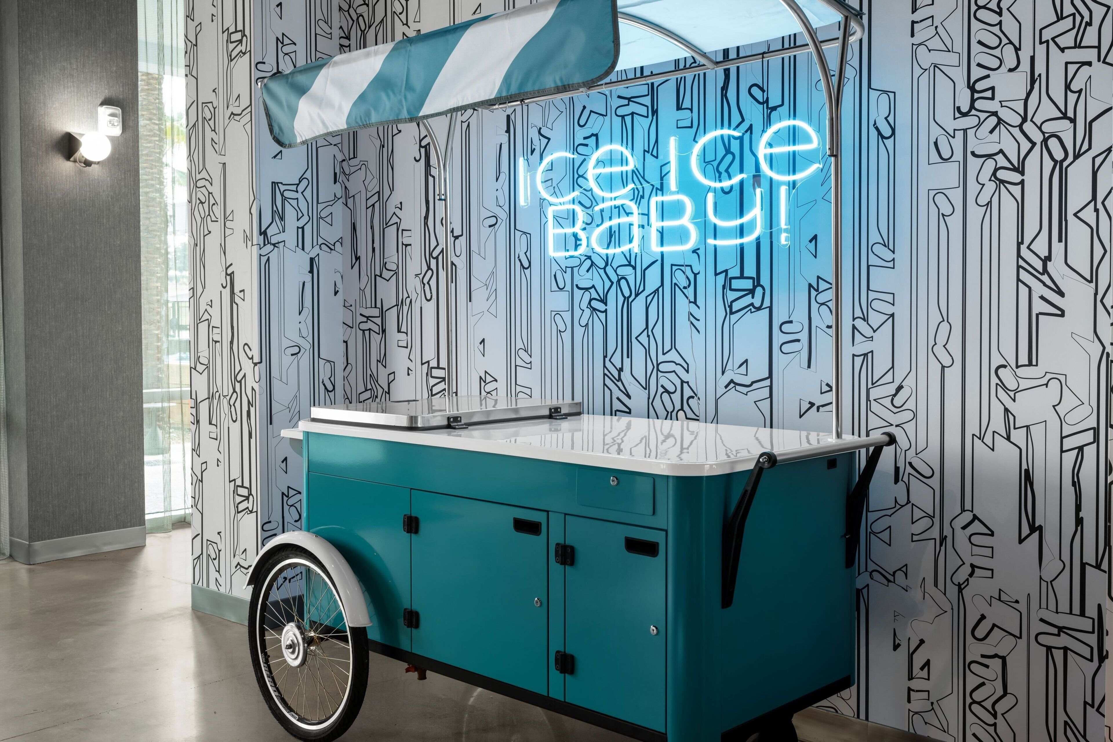 Un chariot de crème glacée turquoise, orné d’un panneau néon sur lequel on peut lire « Ice Baby ! », est placé devant un mur orné d’un motif abstrait en noir et blanc.