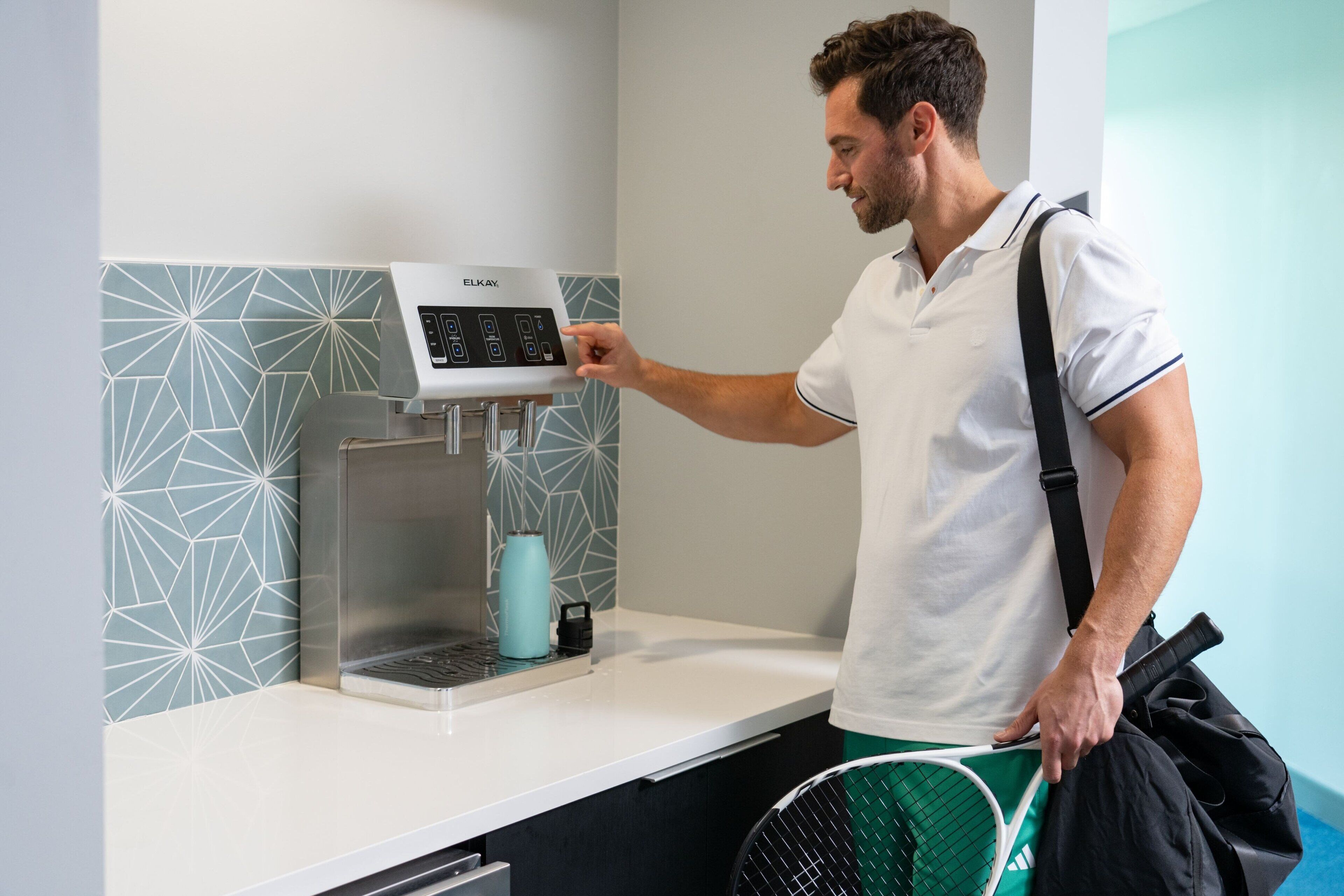 Un homme, vêtu d’un polo blanc et d’un pantalon noir, se tient à côté d’une machine à café dotée d’un écran numérique et tient une raquette de tennis.