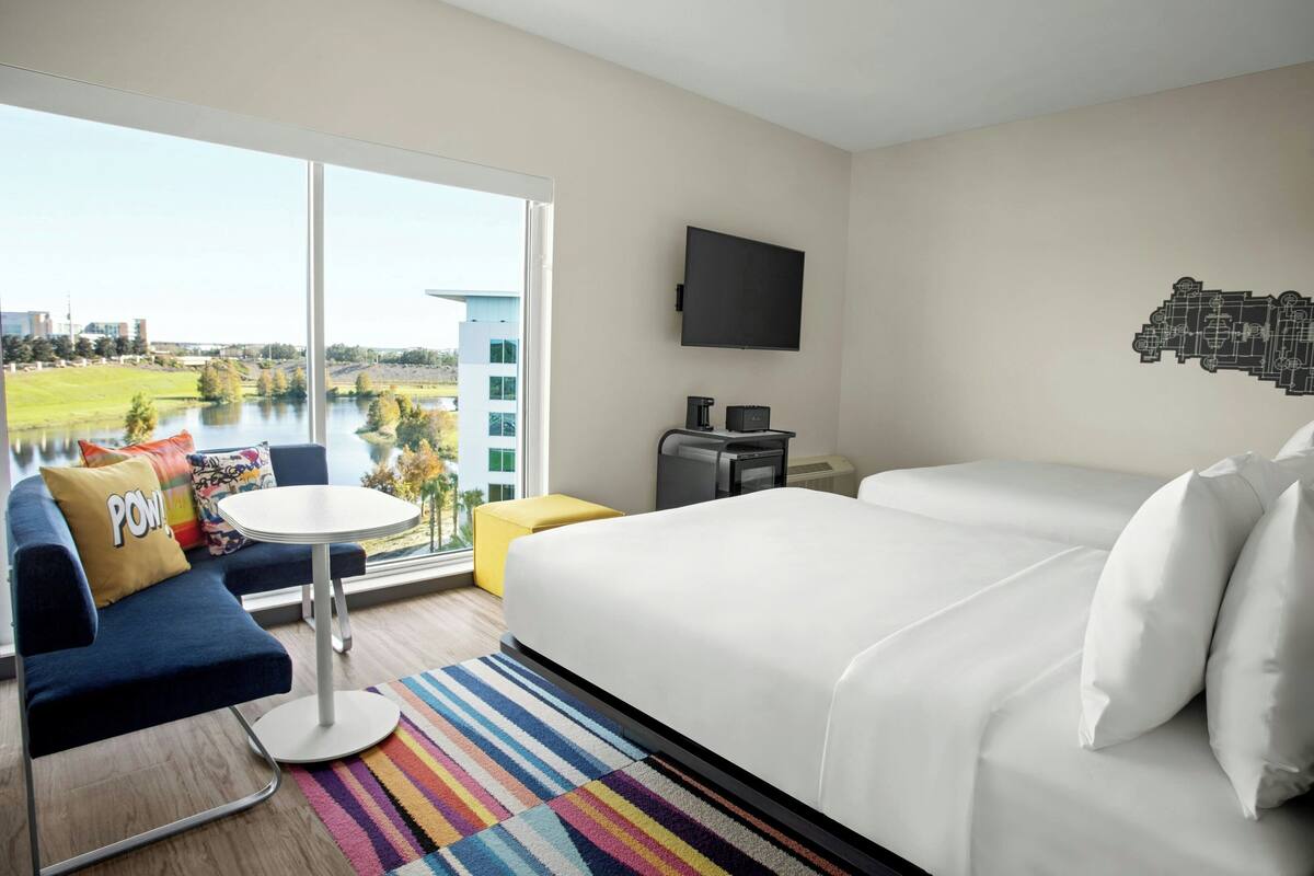 Habitación de hotel con una cama grande, un sofá, una mesita, un televisor y vistas a un lago y edificios.