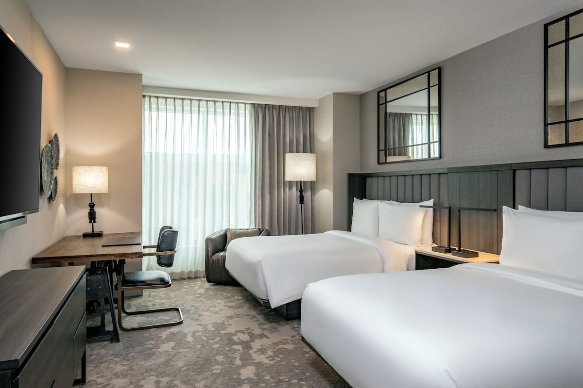 Une chambre d’hôtel avec deux lits, un bureau, une chaise, une télévision et une grande fenêtre avec des rideaux.