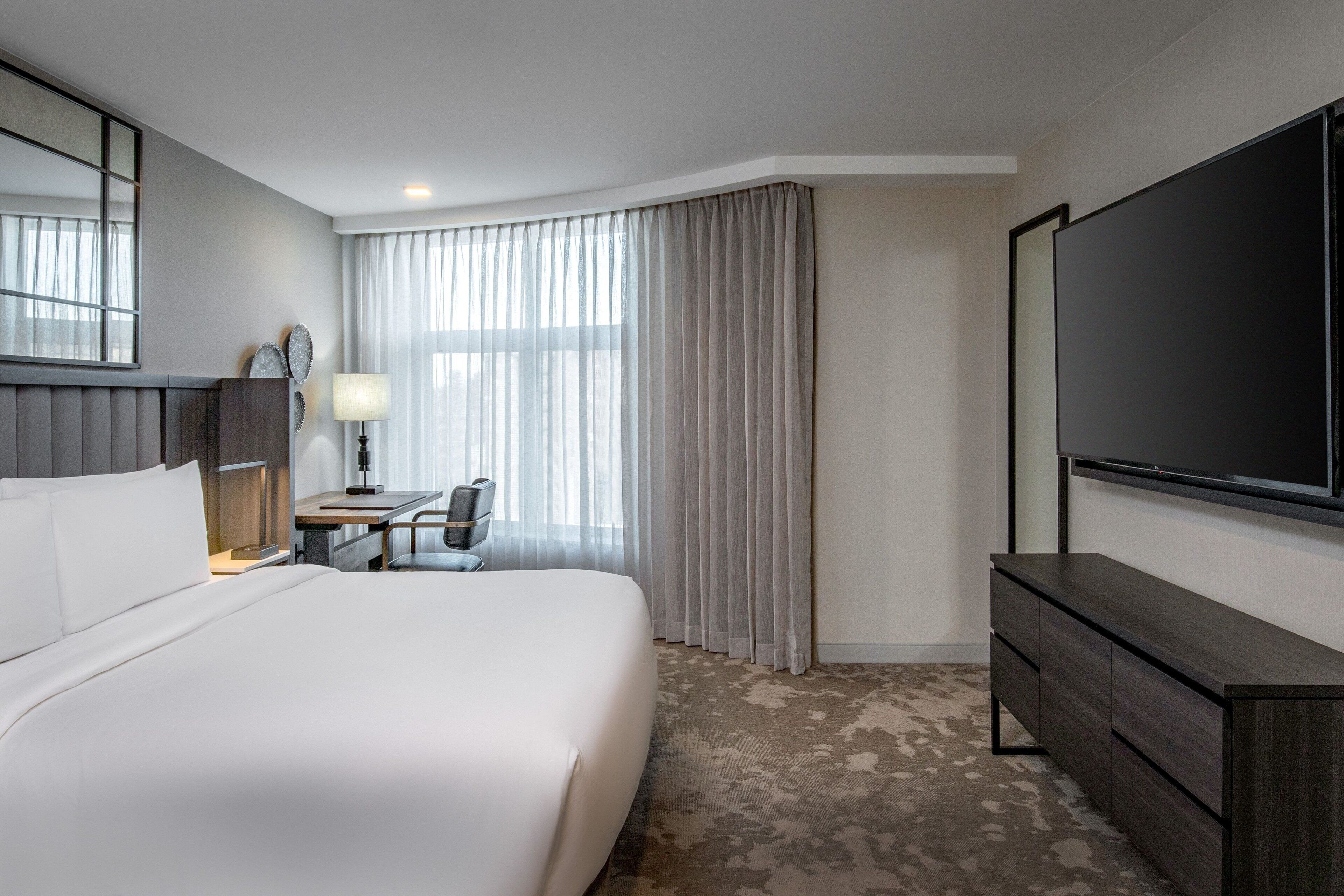 Quarto de hotel com uma cama grande, uma escrivaninha com cadeira, televisão de tela plana, cômoda e uma janela ampla com cortinas.
