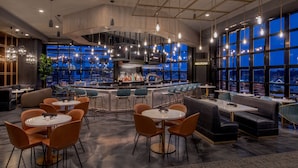 A modern bar area with round tables and orange chairs, a bar counter with a variety of bottles, and large windows offering a view of the city at night. - The Artisan At Tuscan Village, Salem, A Tribute Portfolio Hotel (Salem)