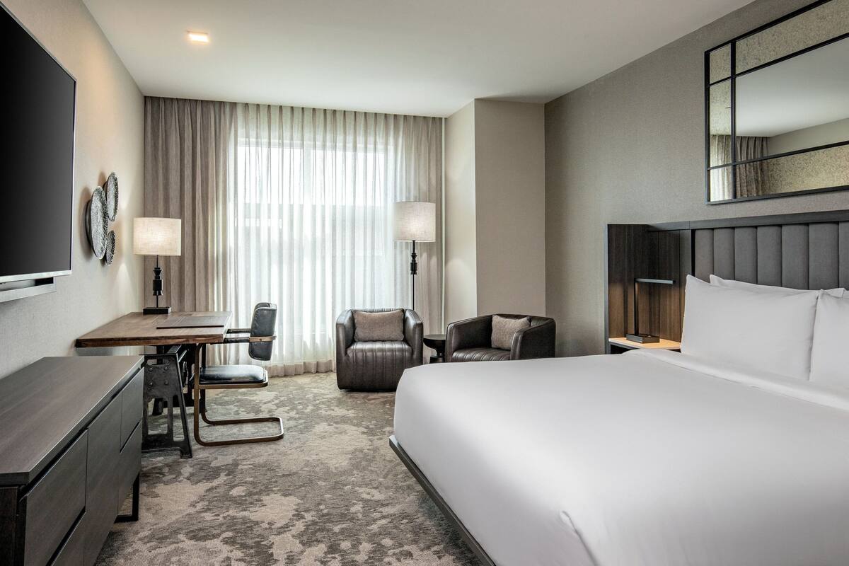 Une chambre d’hôtel avec un grand lit, un bureau avec une chaise, deux fauteuils, une télévision et une grande fenêtre avec des rideaux.
