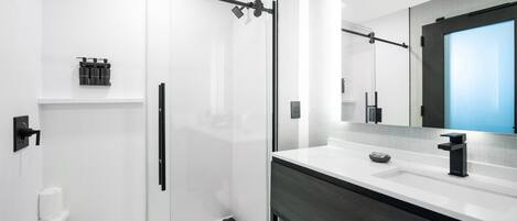 Uma casa de banho moderna com cabine de duche em vidro, lavatório branco com bancada escura e um espelho grande acima do lavatório.