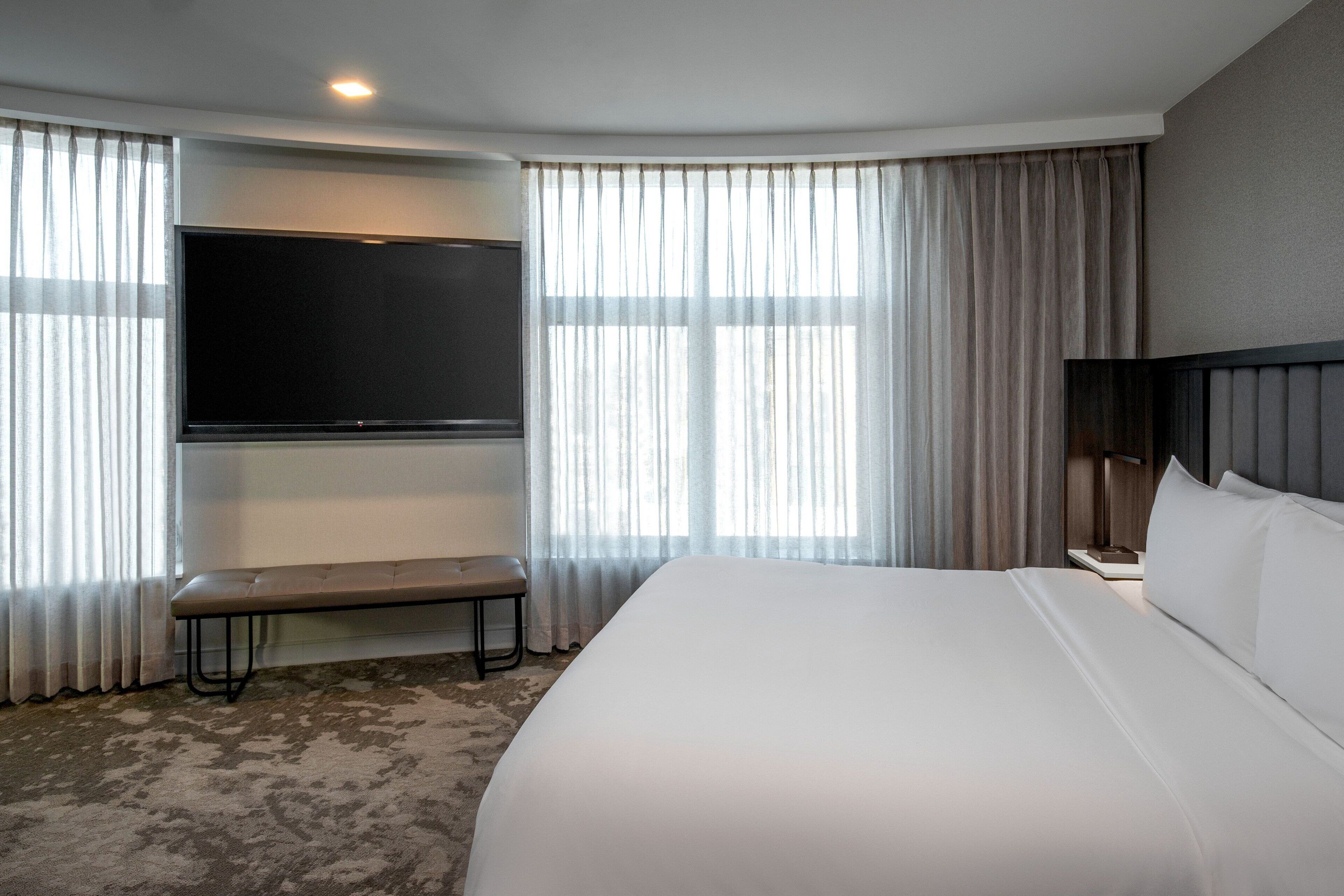 A hotel room with a large bed, a bench, a flat-screen TV mounted on the wall, and a nightstand with a lamp.