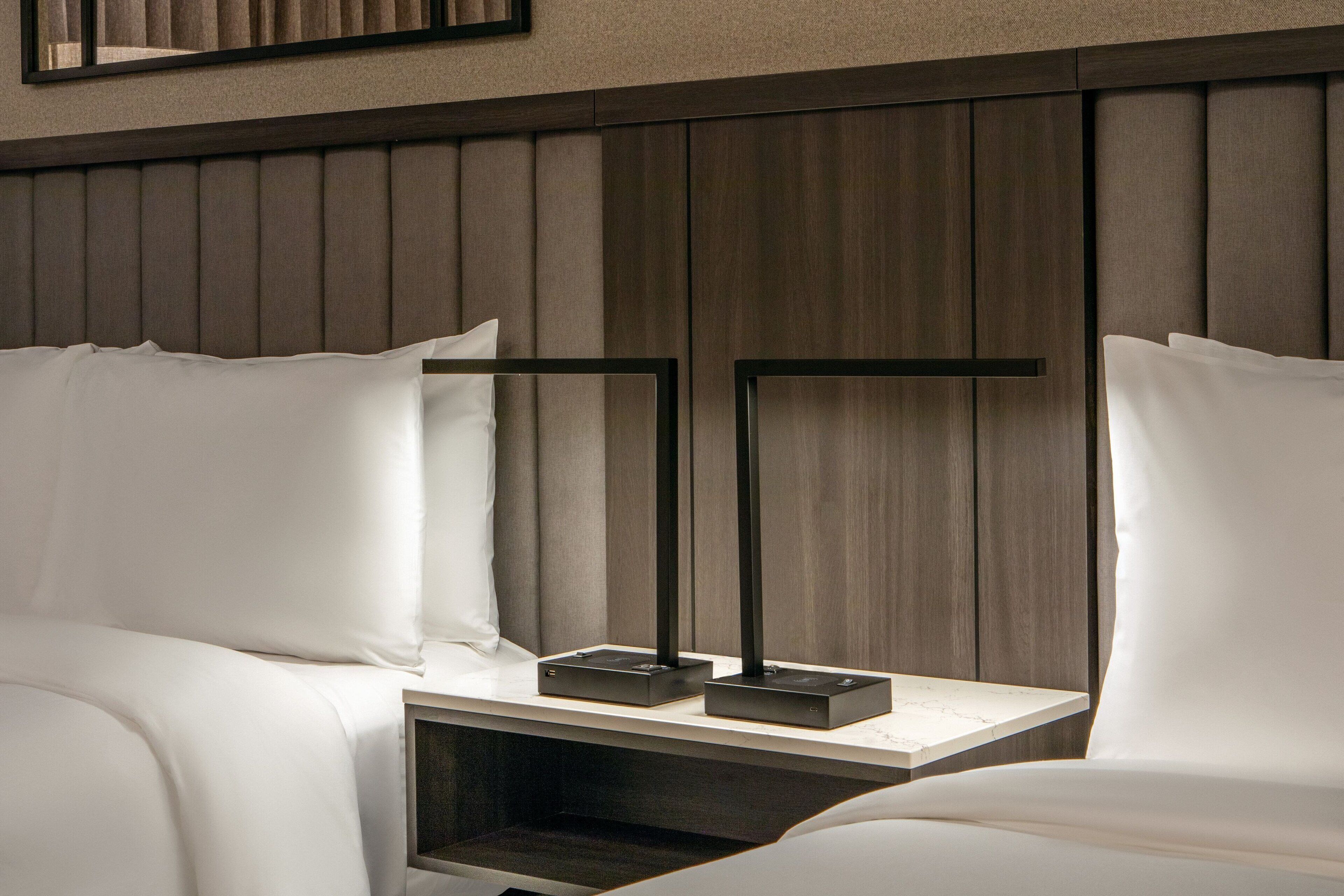 A hotel room with a wooden headboard, two bedside tables with black stands, and white bedding.