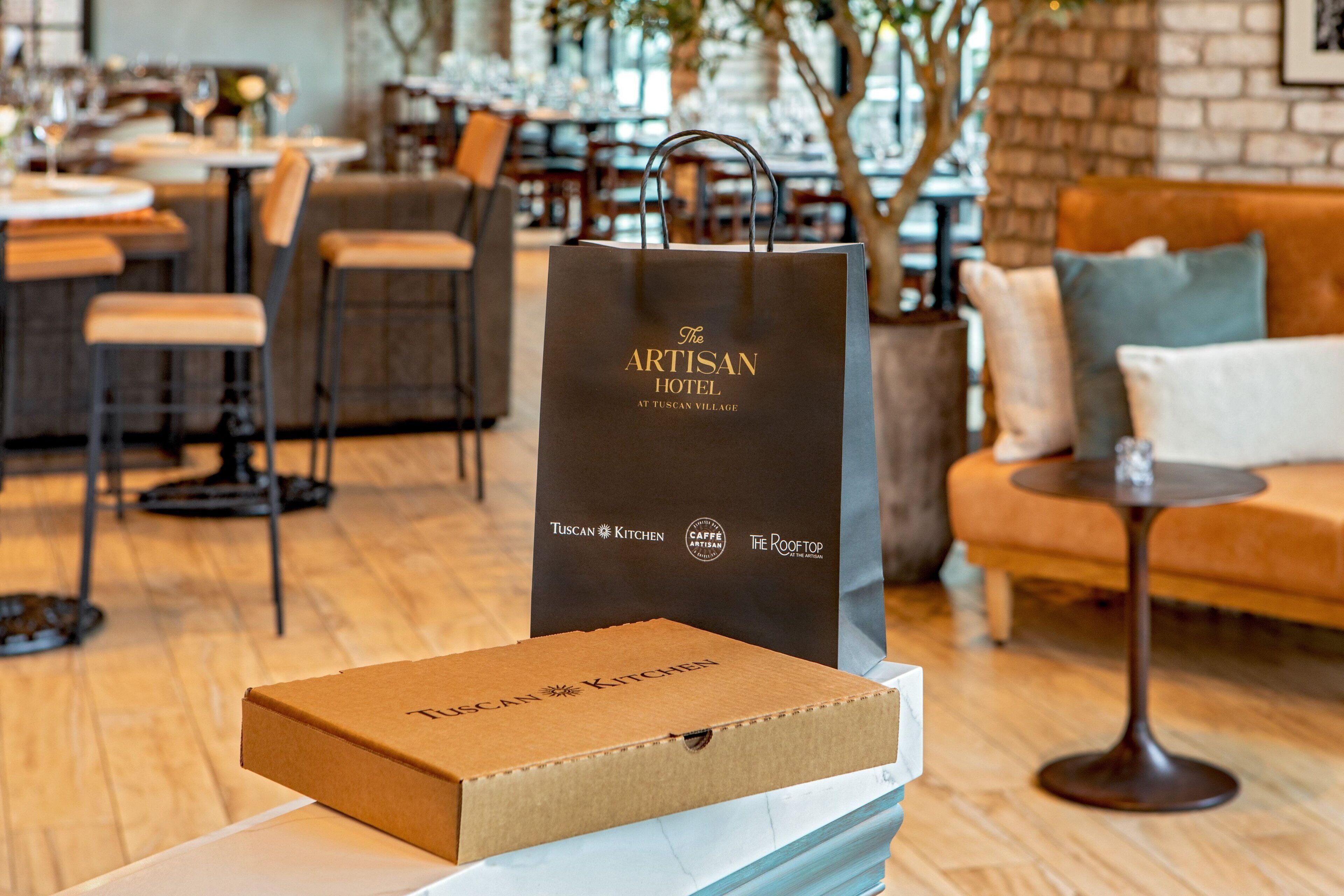 A restaurant interior with wooden flooring, tables, chairs, and a sofa. A brown box labeled "Tuscan Kitchen" and a black bag with "The Artisan Hotel" logo are visible.