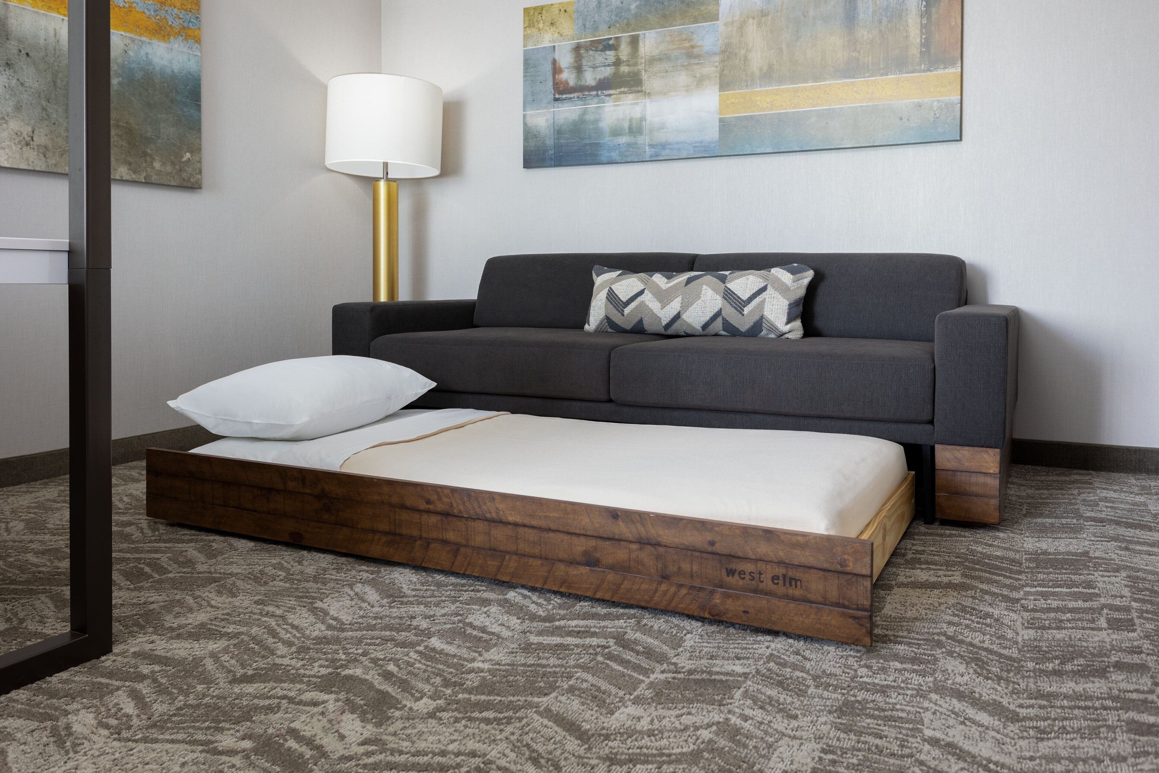 A modern room with a grey sofa, a wooden platform bed, a white pillow, a lamp, and abstract art on the wall.