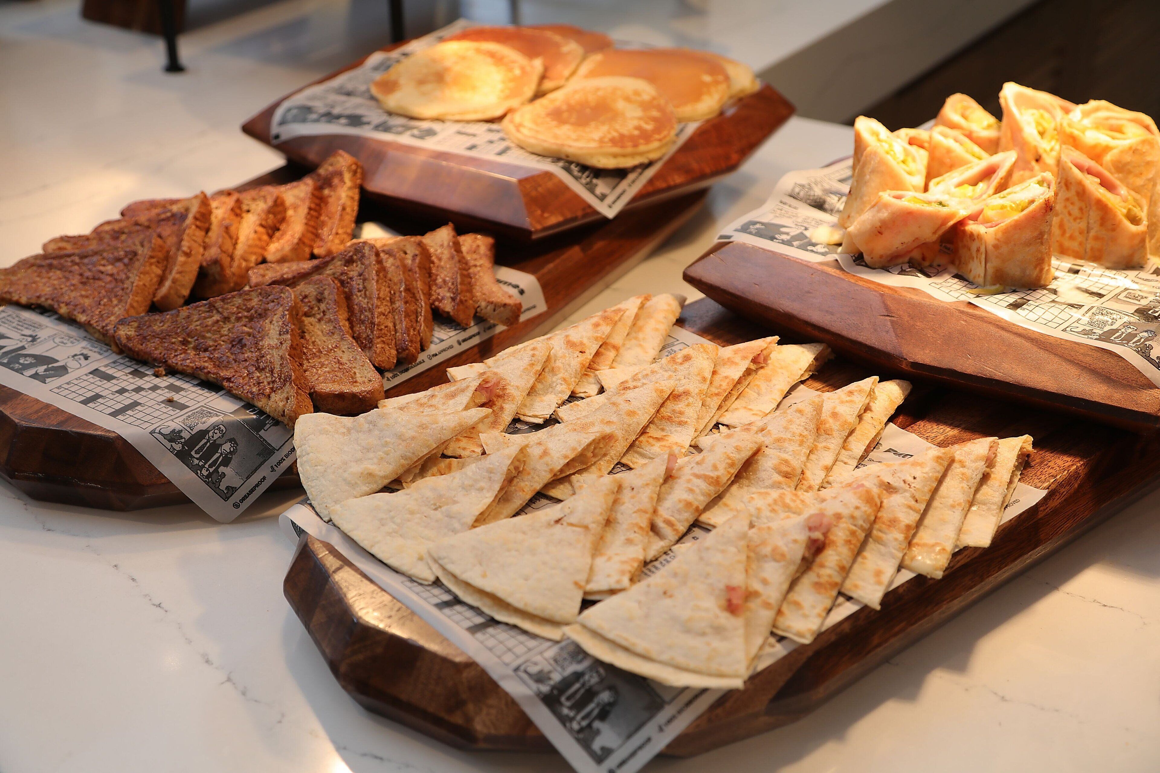 Variedad de alimentos para el desayuno, como tostadas, panqueques y pasteles, sobre bandejas de madera.