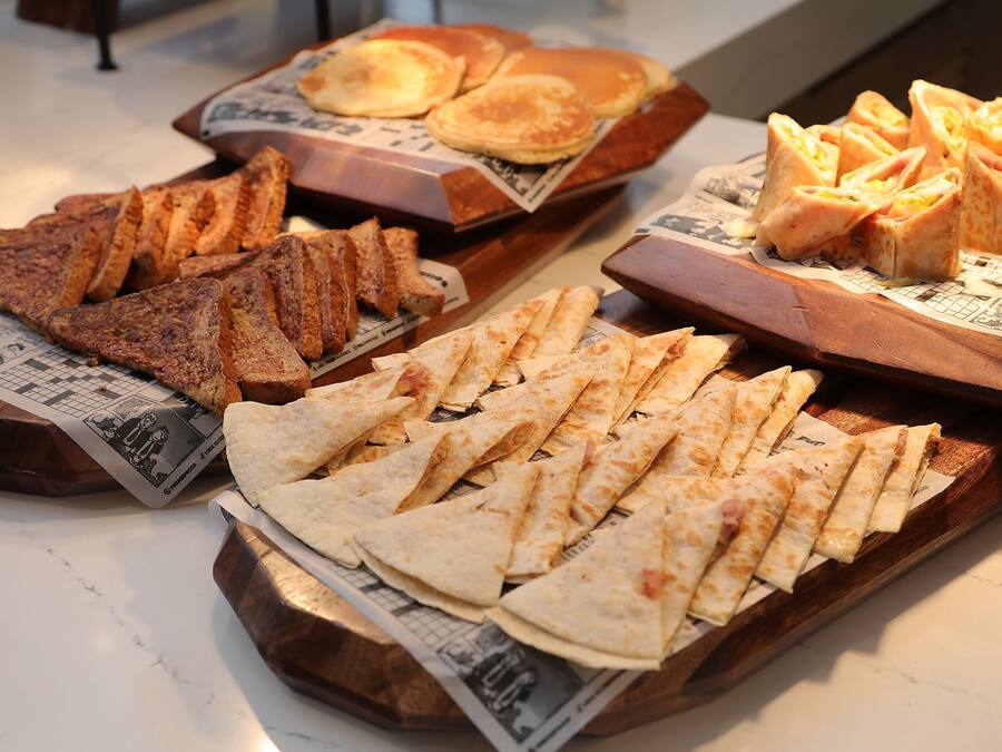 Variedad de alimentos para el desayuno, como tostadas, panqueques y pasteles, sobre bandejas de madera.
