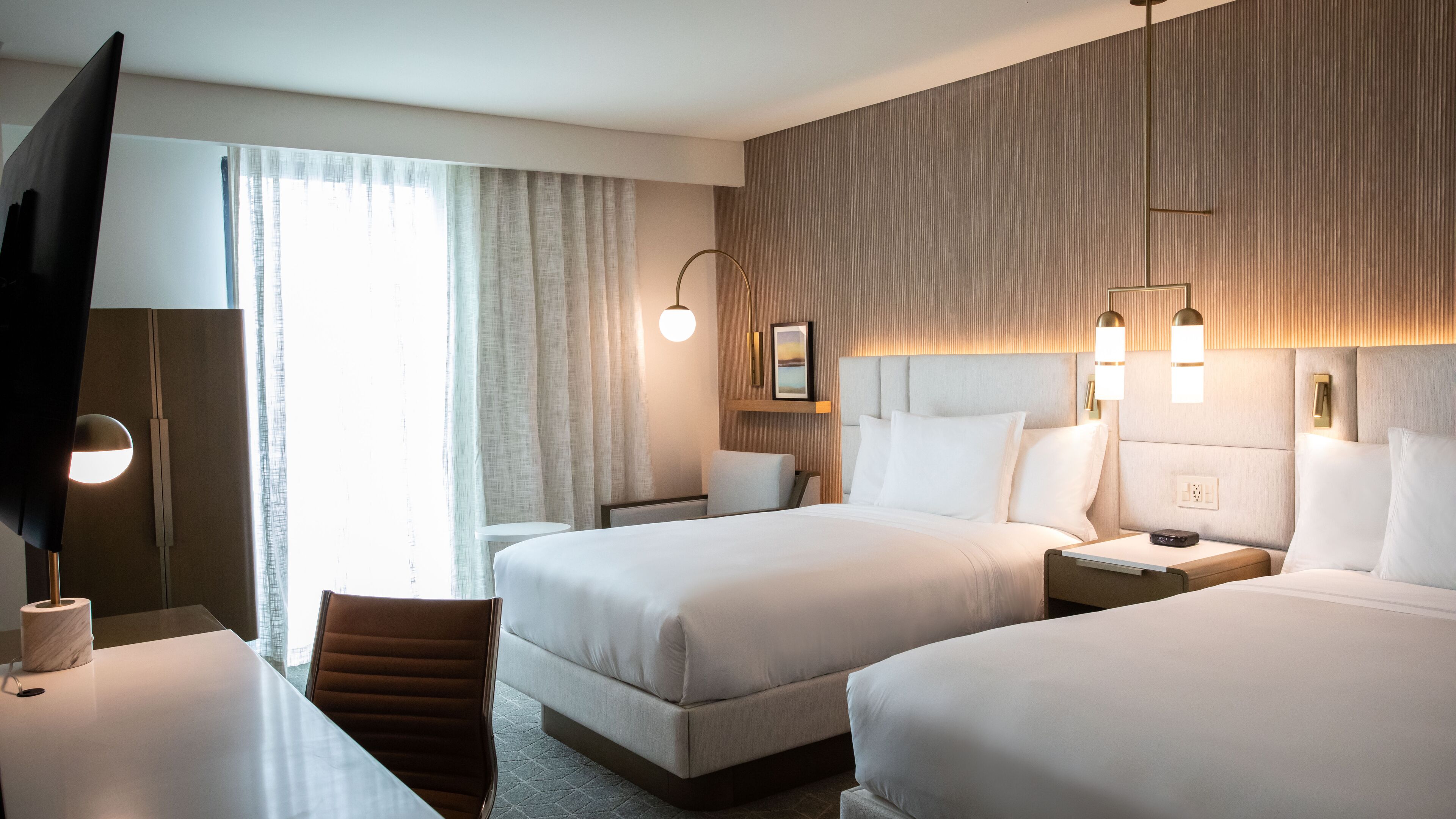 Habitación de hotel con dos camas, un escritorio, una silla, un televisor y un ventanal con cortinas.