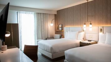 Habitación de hotel con dos camas, un escritorio, una silla, un televisor y un ventanal con cortinas.