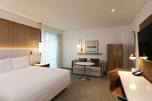 Habitación de hotel con una cama grande, un sofá, una pequeña mesa redonda y un escritorio con silla. Se aprecia un cuadro en la pared, un espejo y un televisor.