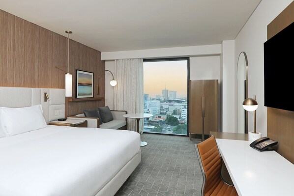 Habitación de hotel con una cama grande, un escritorio, una silla, una mesita, una lámpara, un teléfono y vistas a la ciudad a través de un amplio ventanal.