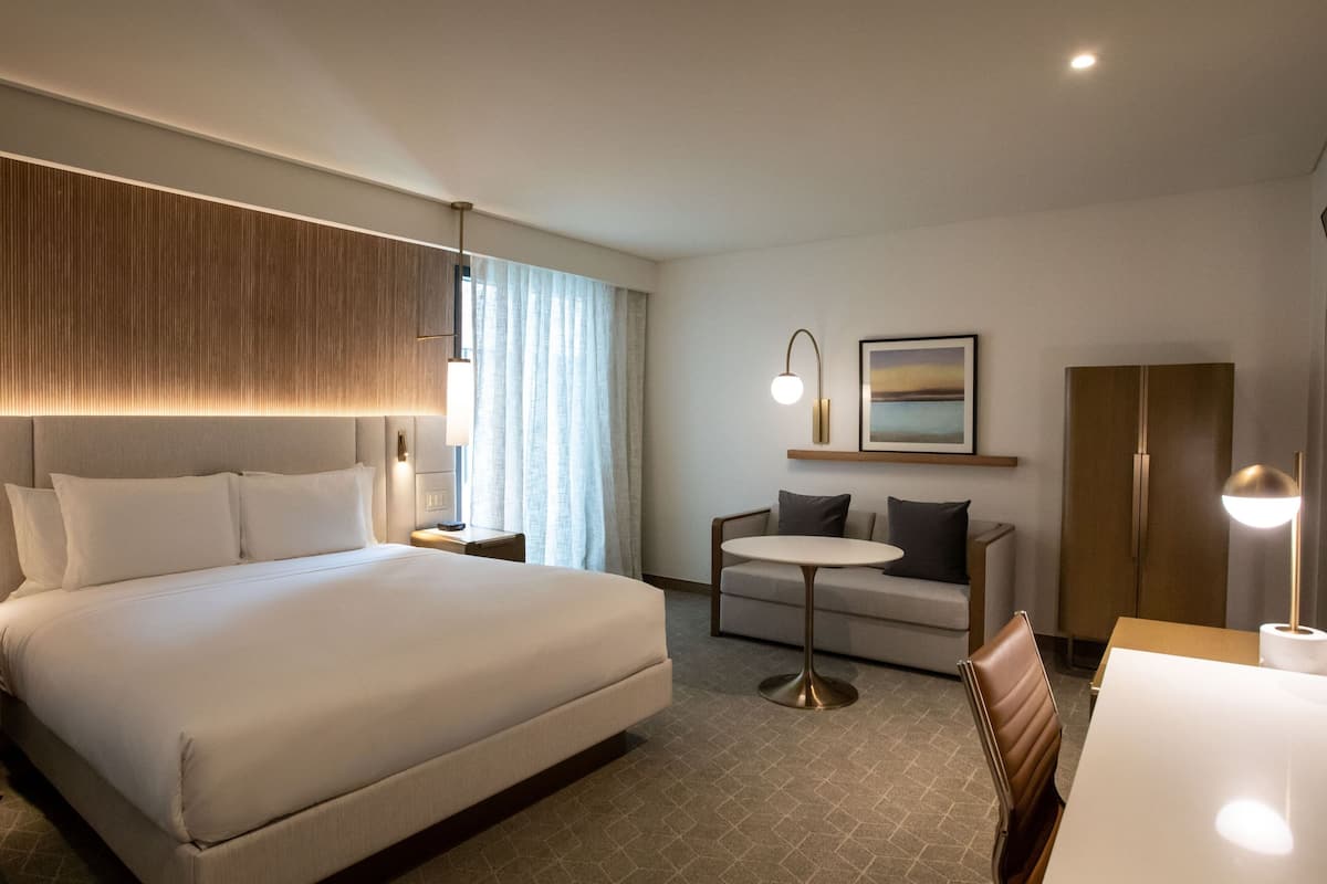 Una habitació d'hotel amb un llit gran, un sofà, una taula rodona petita i un escriptori amb una cadira. Hi ha dues làmpades penjades a la paret i un quadre emmarcat sobre el sofà.
