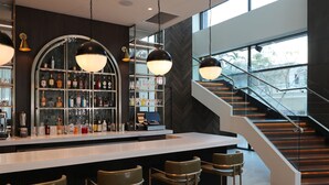 A modern bar area with a white countertop, multiple stools, and a variety of bottles and glasses on shelves. A staircase with glass railings leads to an upper level. - Courtyard By Marriott Santo Domingo Piantini (Santo Domingo)