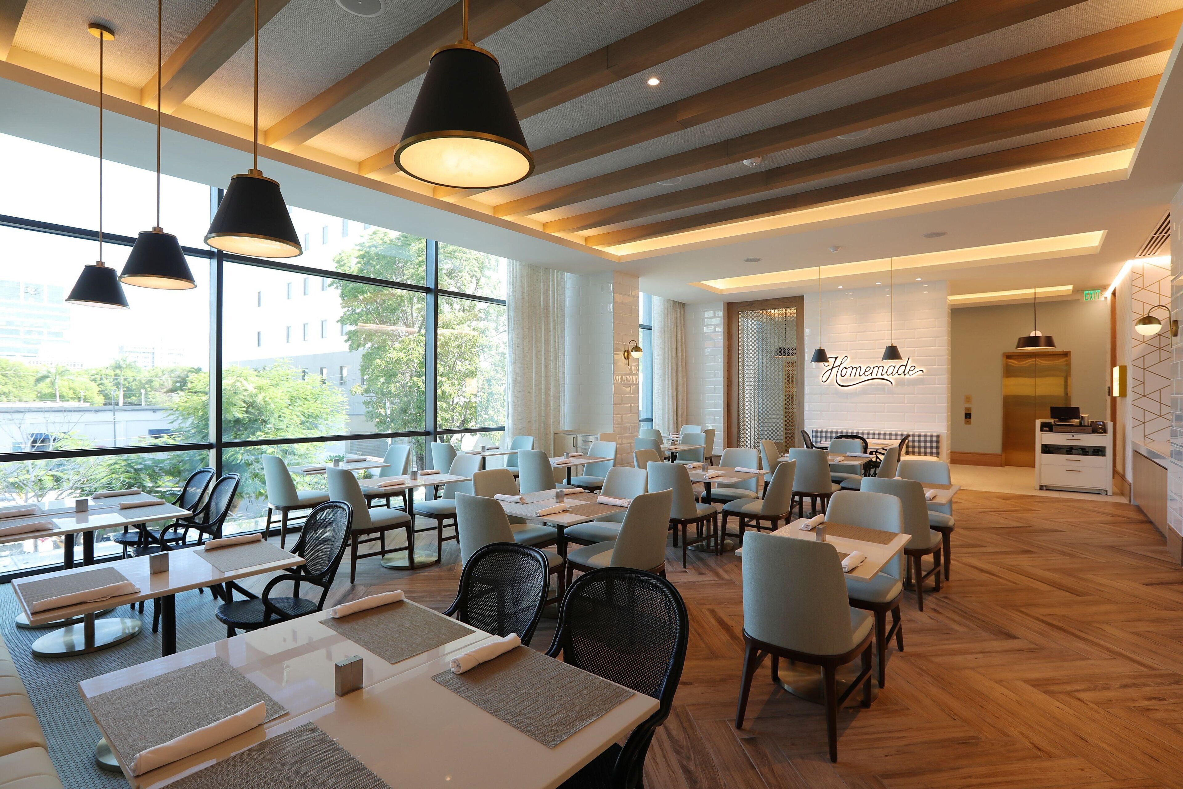Restaurante moderno com grandes janelas, vigas de madeira no teto e luminárias pendentes pretas. Mesas dispostas de forma organizada com cadeiras.