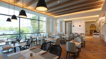 Restaurante moderno com grandes janelas, vigas de madeira no teto e luminárias pendentes pretas. Mesas dispostas de forma organizada com cadeiras.