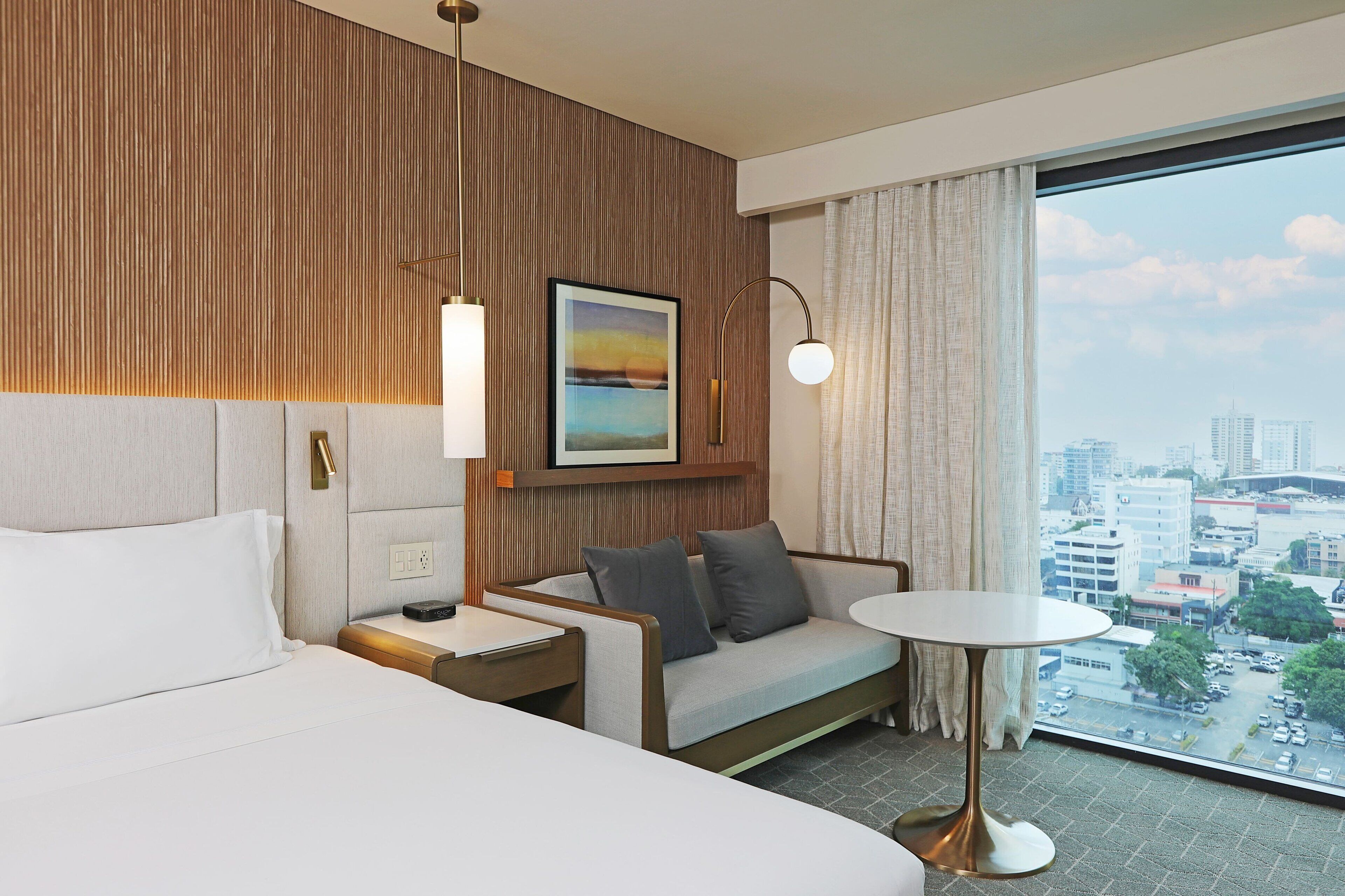 Ein Hotelzimmer mit Bett, Sofa, einem kleinen Tisch und einem großen Fenster mit Blick auf die Stadt.