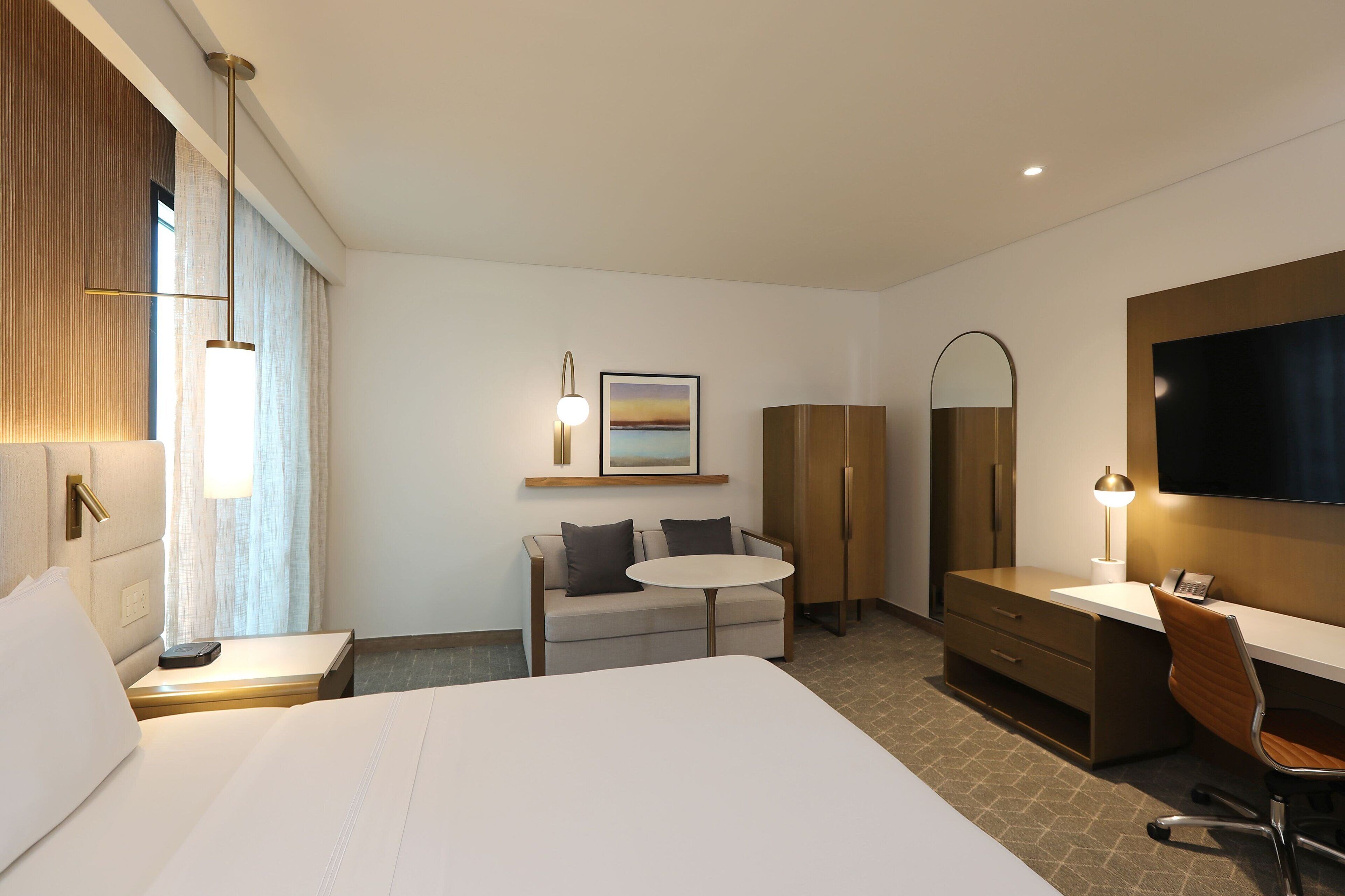 Habitación de hotel con una cama grande, un escritorio con silla, una mesita redonda pequeña, un sofá, un armario, un televisor y un cuadro en la pared.