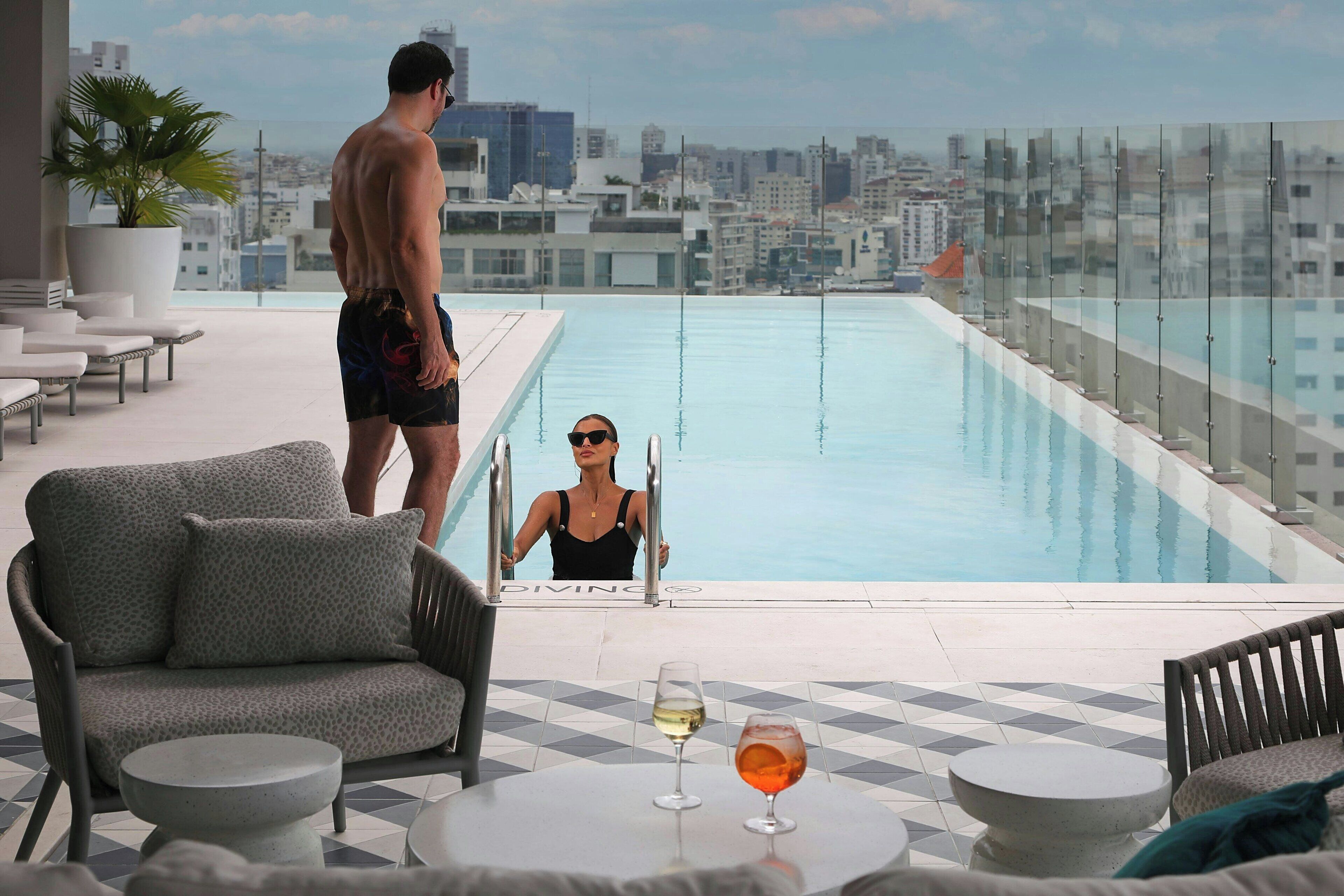Zona de piscina en la azotea con vistas a la ciudad, sillas de descanso y una mesa con bebidas.
