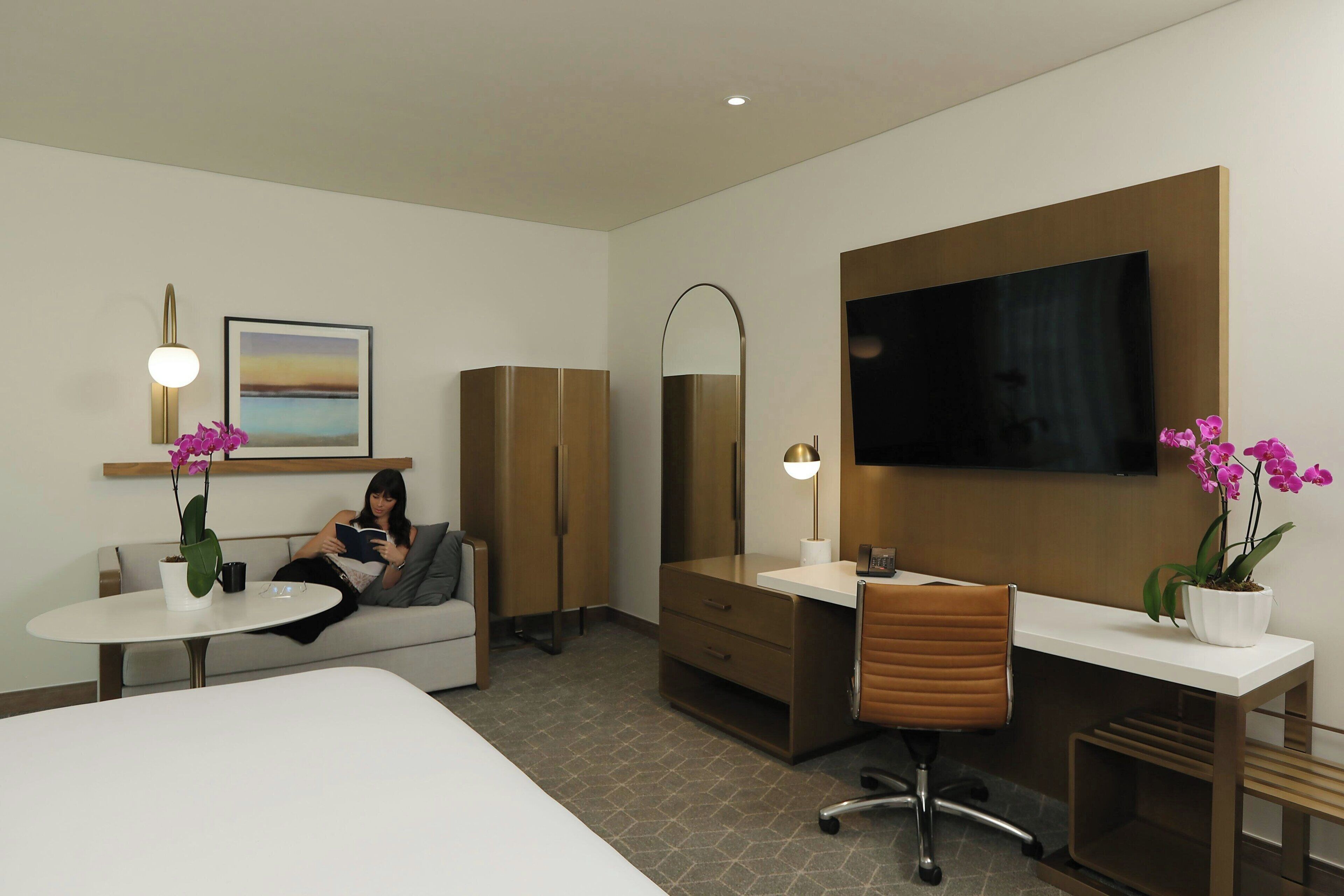 Een hotelkamer met een bed, een bureau, een stoel, een televisie, een bank, een salontafel en twee potplanten.