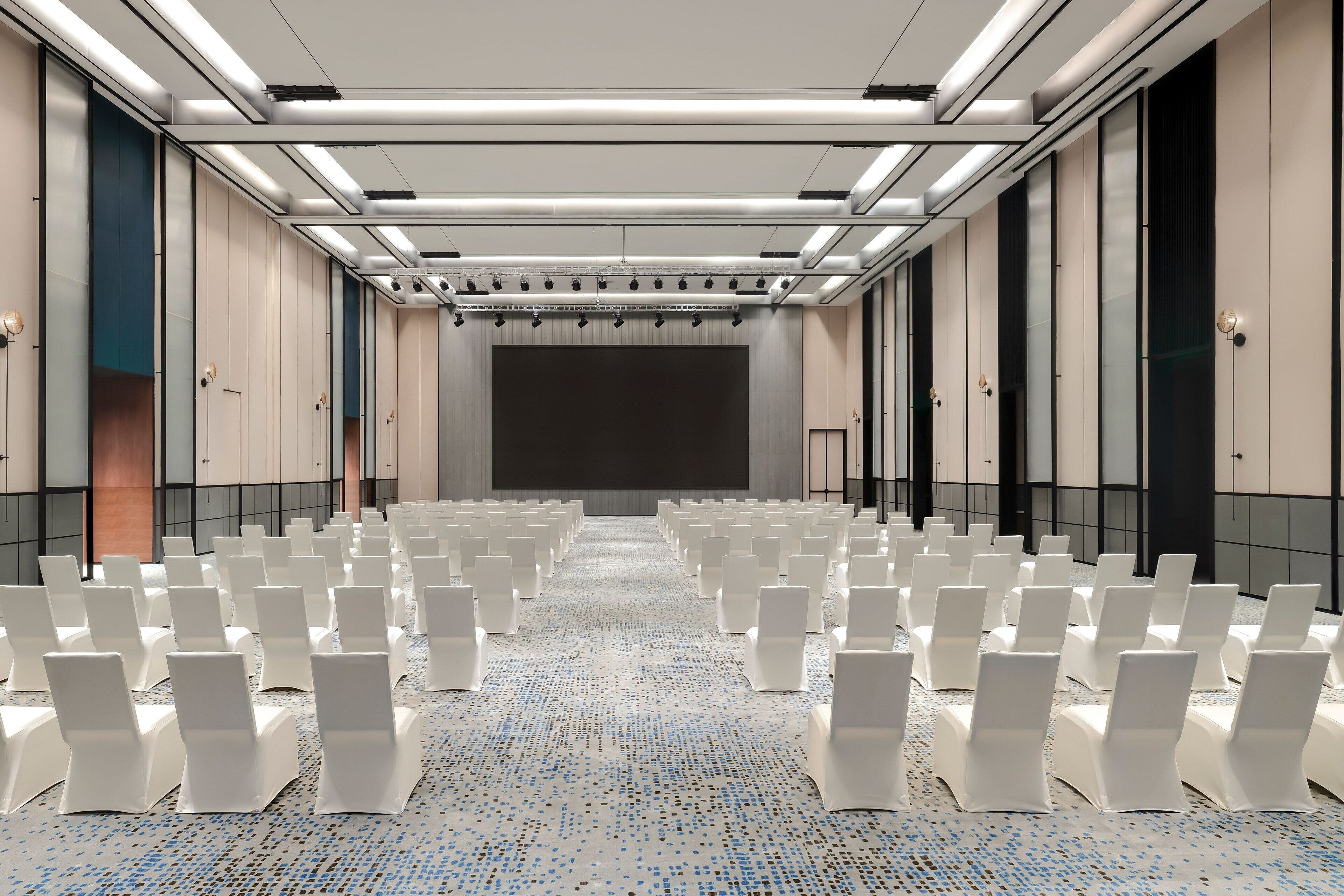 A conference room with rows of white chairs, a large screen at the front, and a patterned carpet.