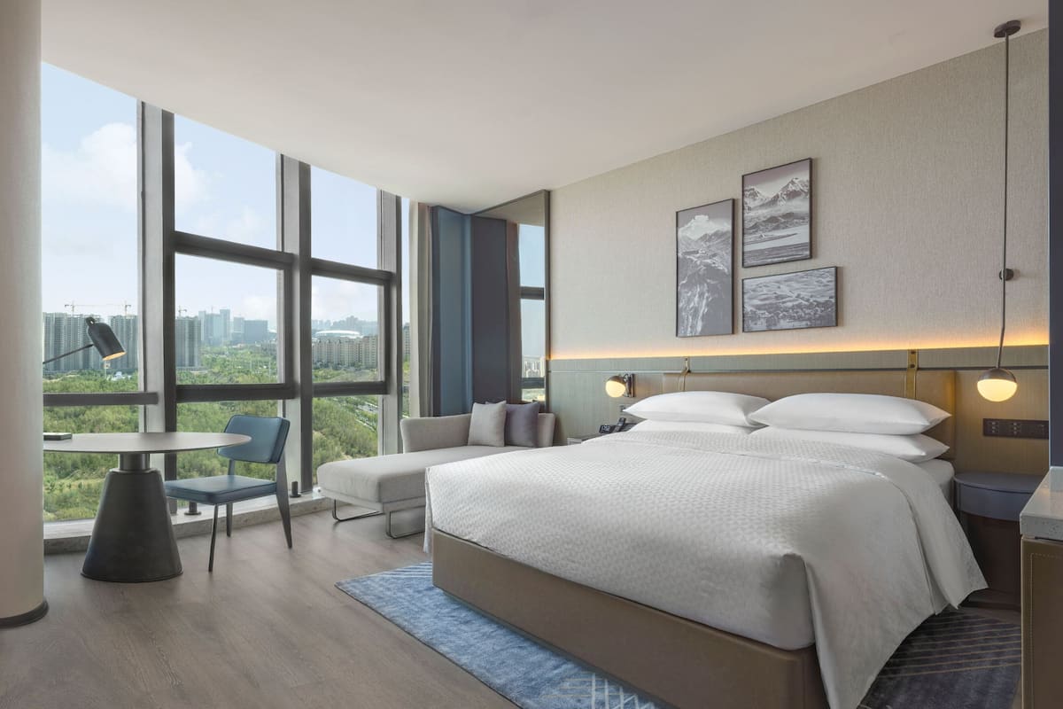 A modern hotel room with a large bed, a seating area with a table and chairs, and large windows offering a city view.
