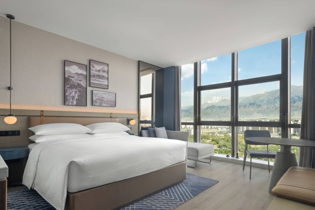 A modern hotel room with a large bed, a desk, and a view of mountains through large windows.