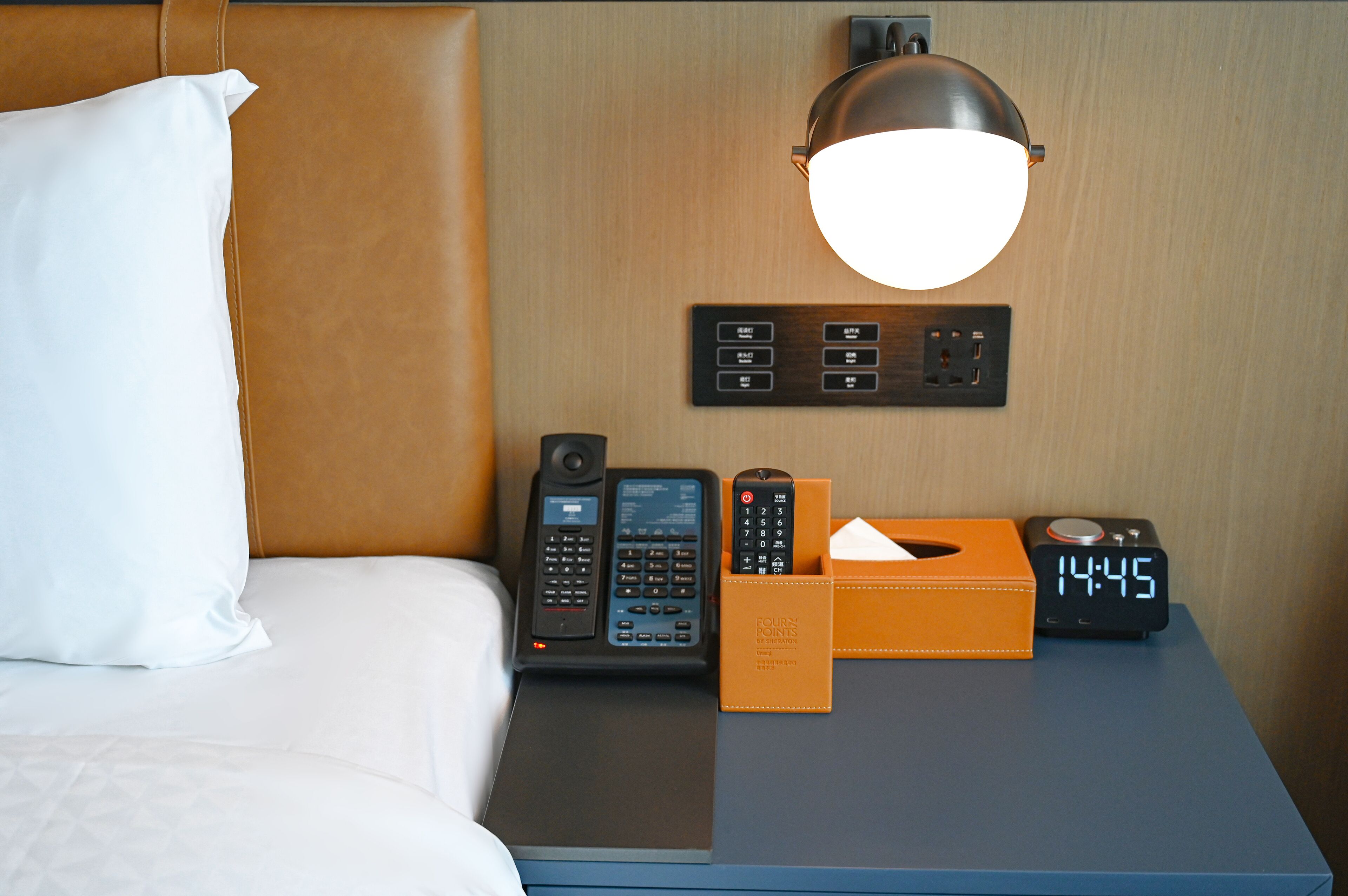 A hotel room with a bed, a nightstand, a lamp, a phone, a clock, and a box of tissues.