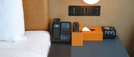 A hotel room with a bed, a nightstand, a lamp, a phone, a clock, and a box of tissues.
