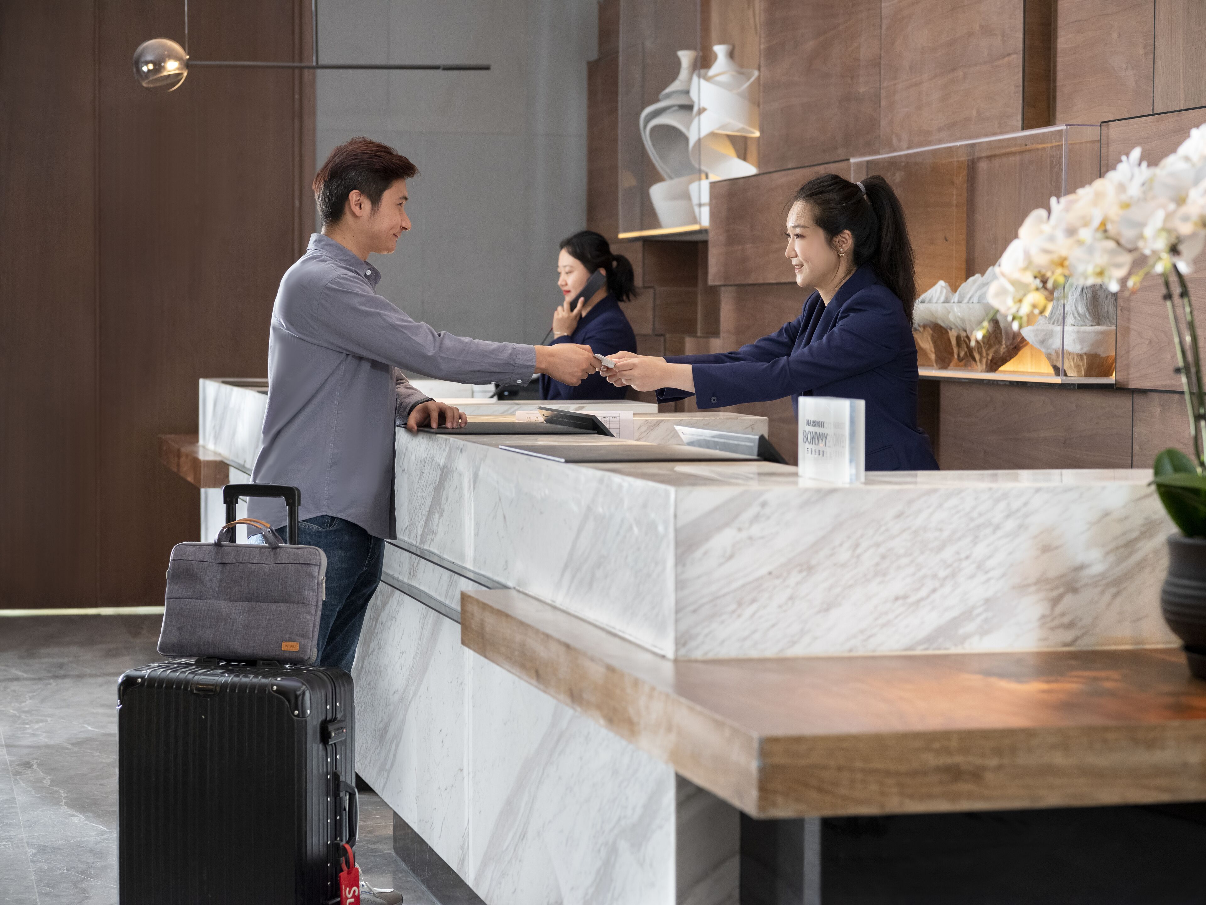 A hotel reception desk with marble countertops, a black suitcase, and a vase with white flowers.
