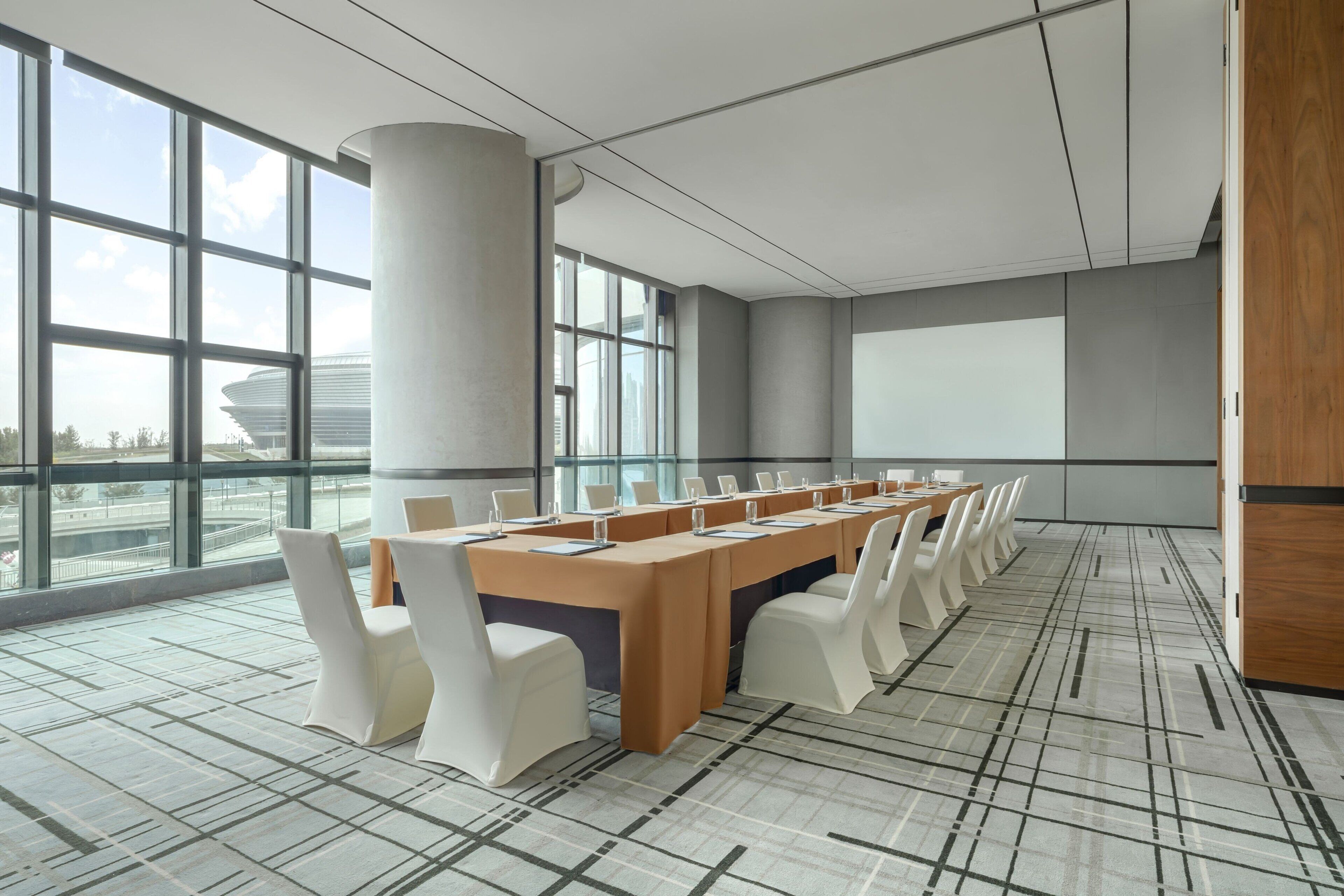 Uma sala de conferências com uma mesa comprida, cadeiras brancas e uma tela grande.