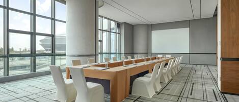 A conference room with a long table, white chairs, and a large screen.