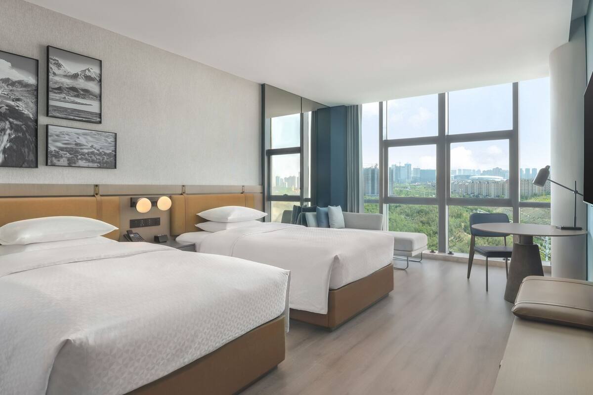 A hotel room with two beds, a desk, and a large window with a city view.