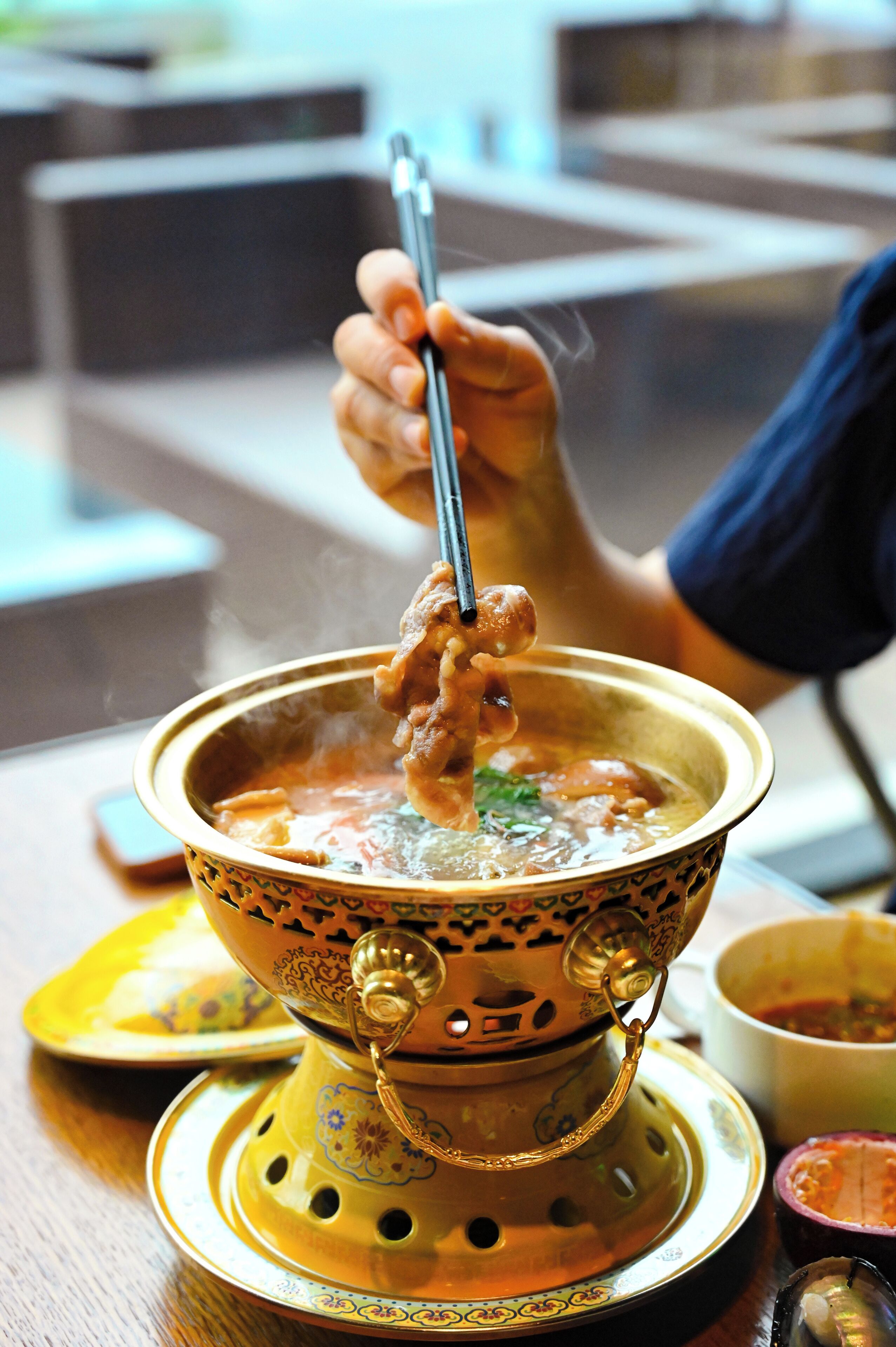 A person holding chopsticks over a bowl of soup, with a decorative teapot and a plate on the table.
