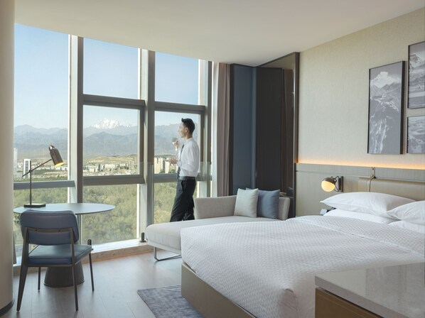A modern hotel room with a large bed, a desk, and a chair. There is a view of mountains through the window. - Four Points By Sheraton Urumqi (Ürümqi)
