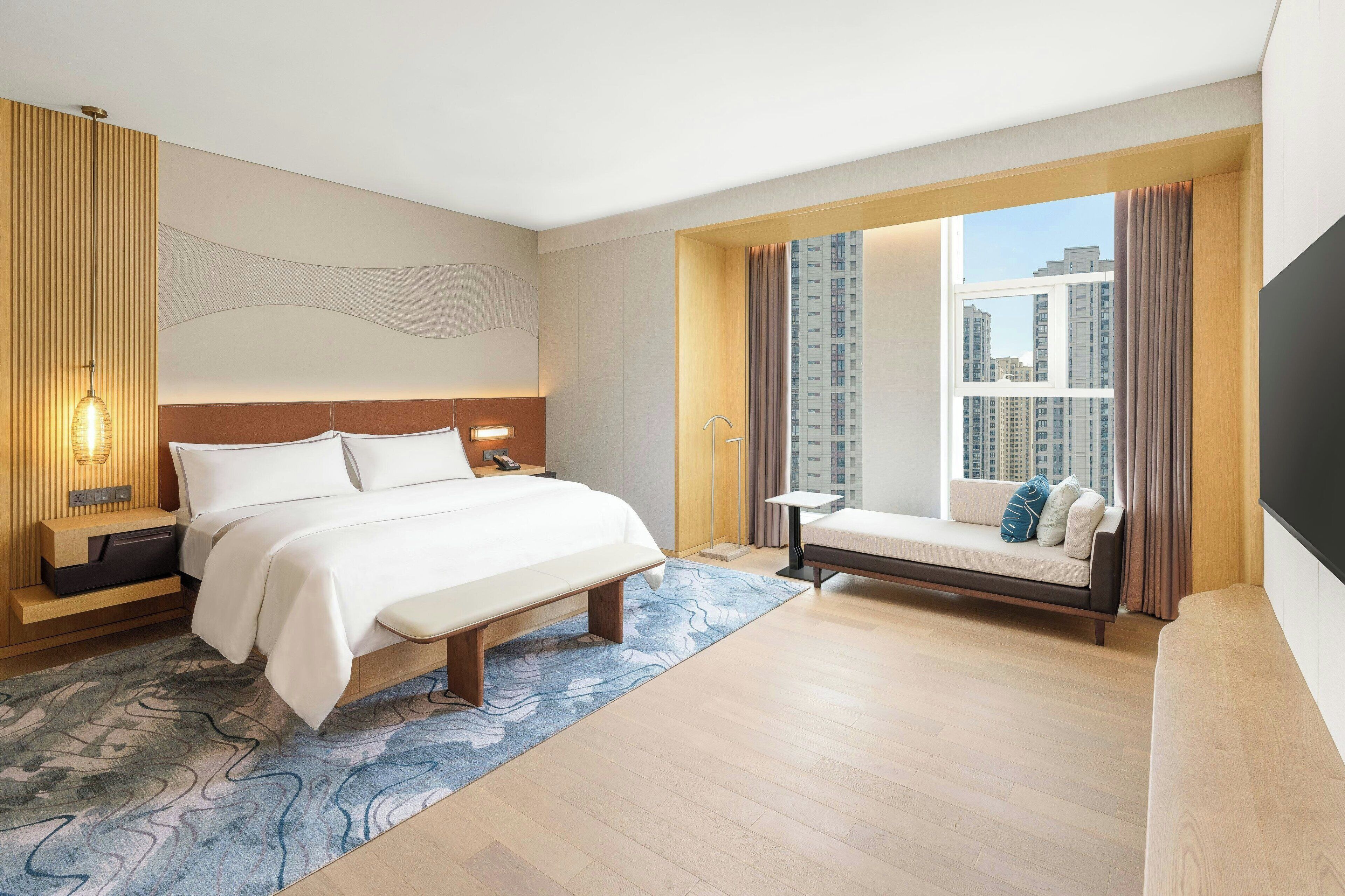 A modern hotel room with a large bed, a sofa, a TV, and a view of high-rise buildings.
