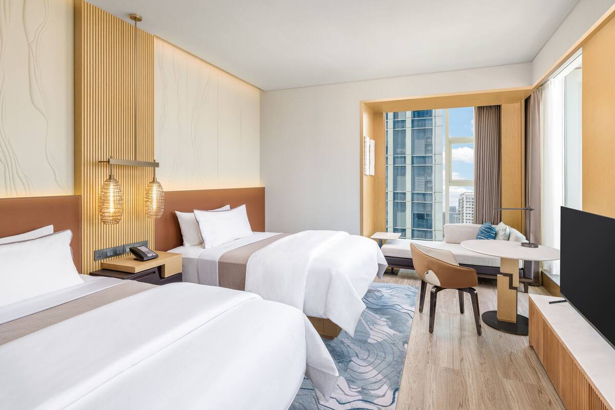 Habitación de hotel con dos camas, un escritorio, una silla, un televisor y un amplio ventanal con vista a la ciudad.