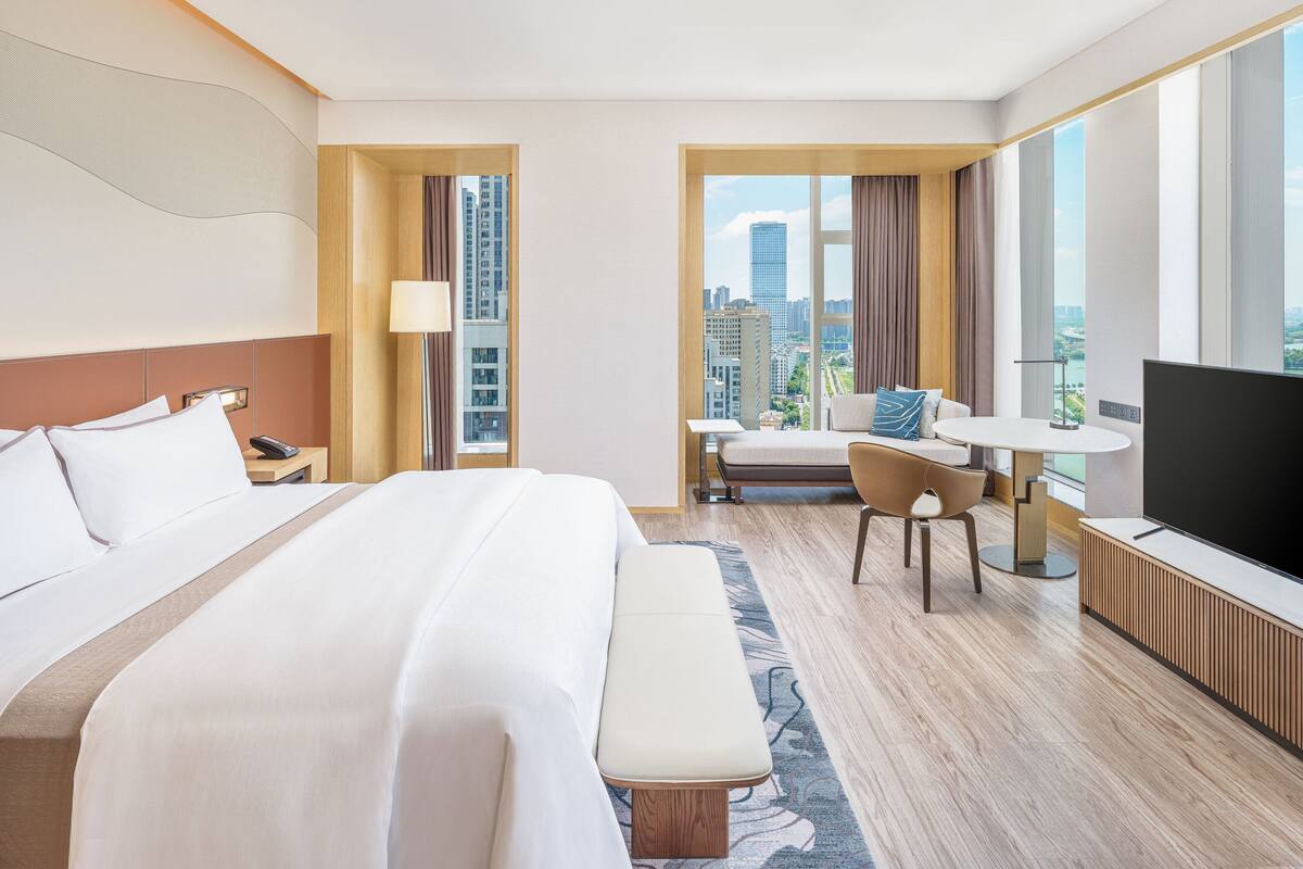 Habitación de hotel con una cama grande, un escritorio con silla, televisor y vista a la ciudad.