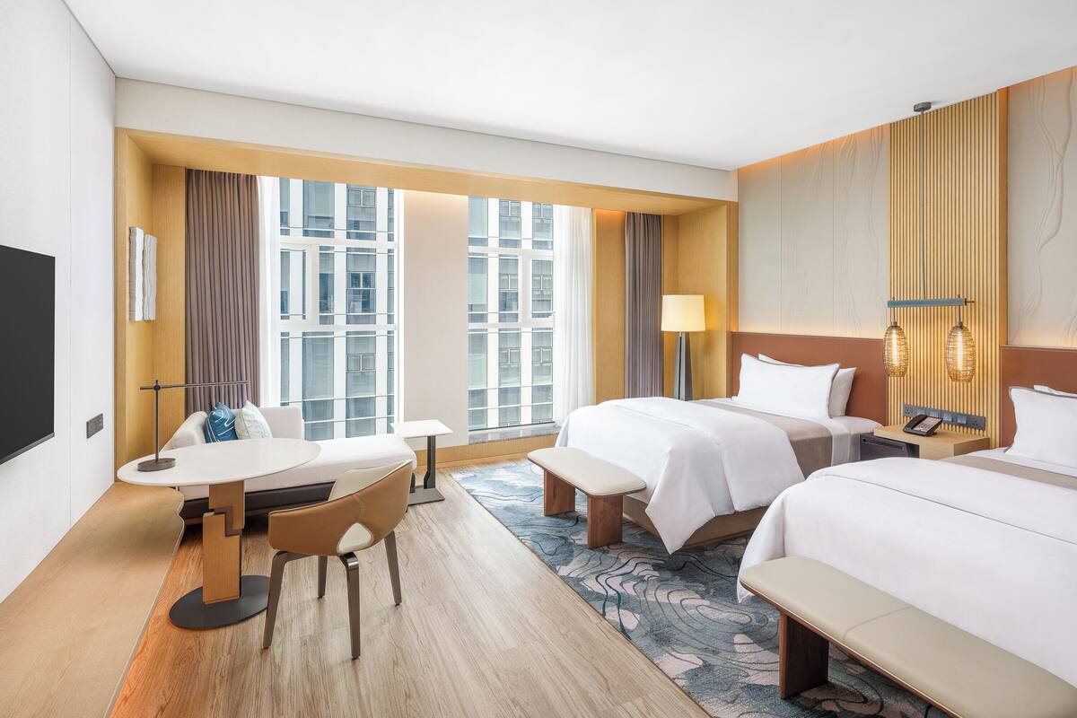 Habitación de hotel con dos camas, un escritorio y una silla. Un ventanal grande ofrece vistas a edificios.