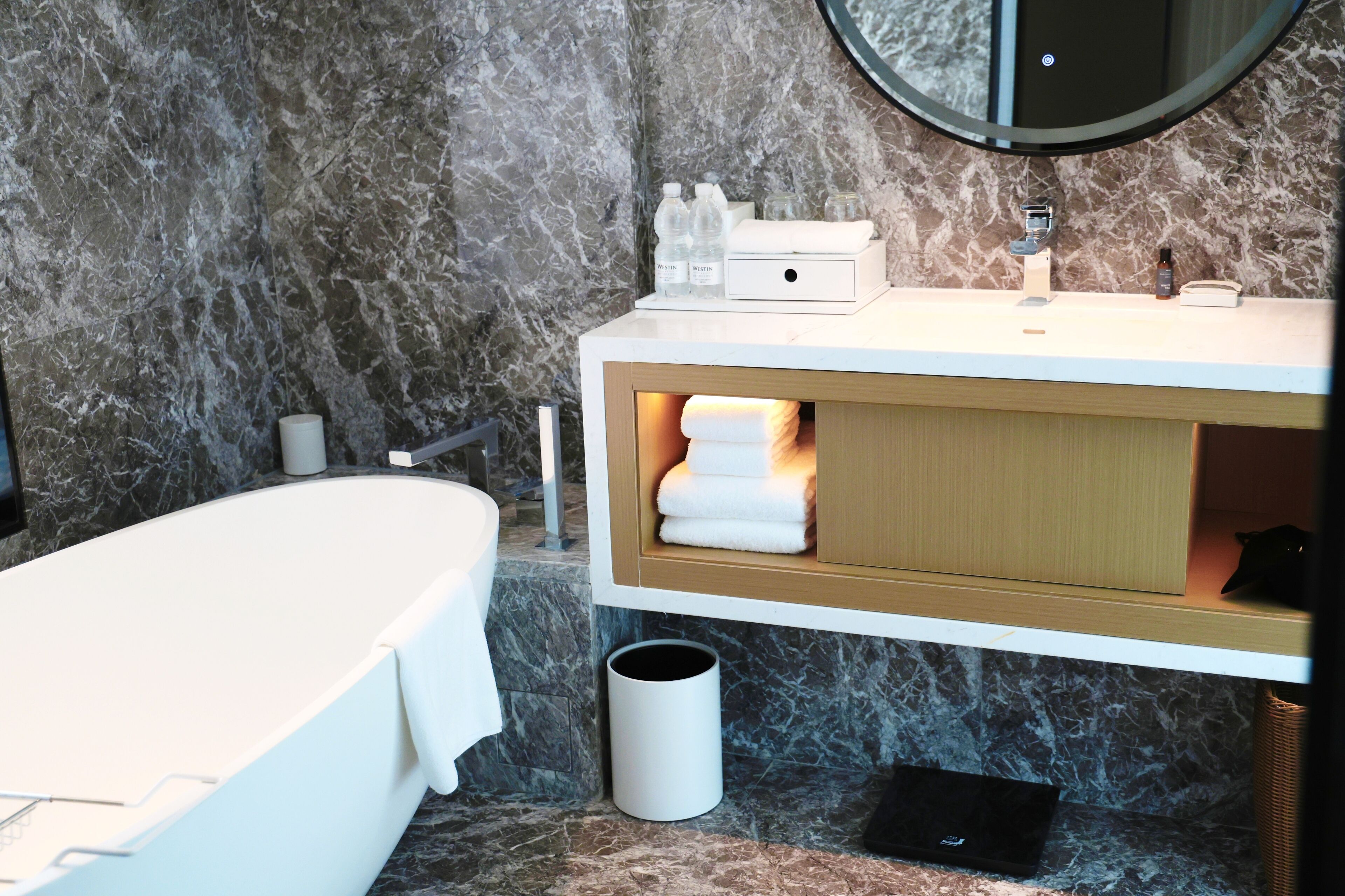 A modern bathroom with a marble wall, a white bathtub, a vanity with a mirror, and a towel rack.